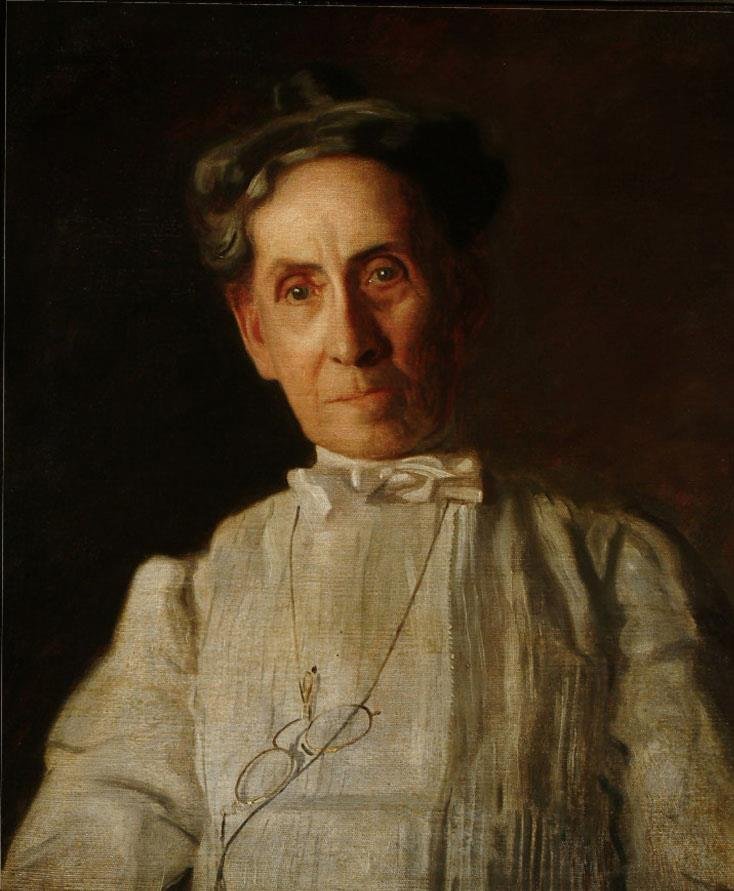 Portrait of an older woman in a white blouse. She wears glasses around her neck on a chain. The woman has her gray hair pulled back into a high bun. She has a wrinkled face with deep set eyes. 