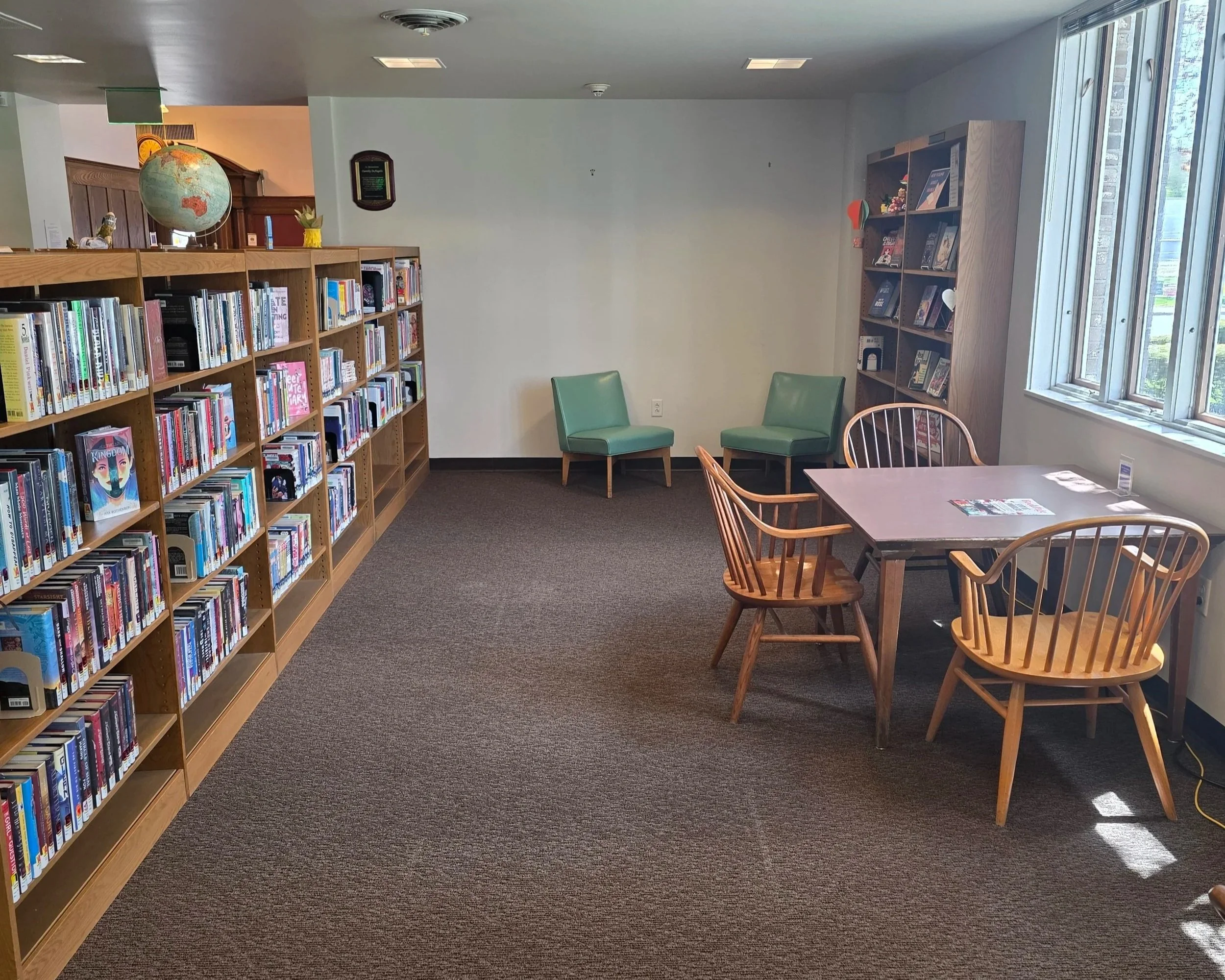 This is a photo of the Teen area at the Arkell Museum and Canajoharie Library; A room created by bookshelf dividers. It has a table with three wooden chairs surrounding it and two comfy lounge chairs parallel to the table.