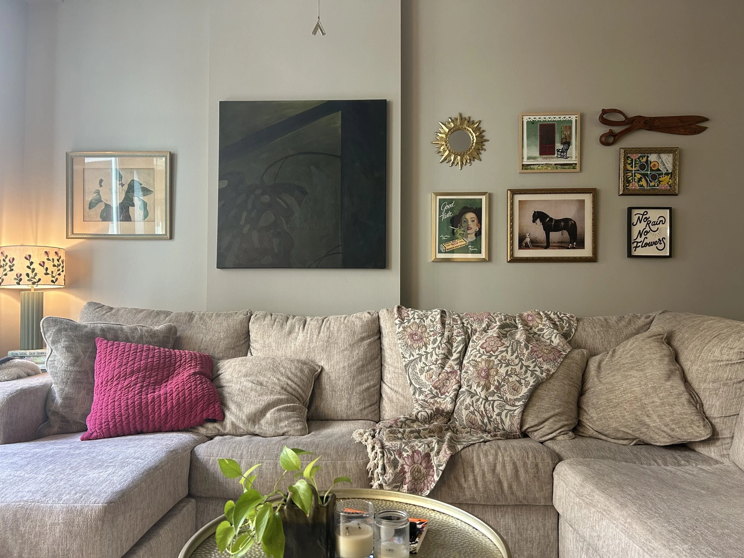 Living room with a beige sectional sofa, colorful pillows, and a coffee table with candles and a plant, wall decor including framed pictures and a small mirror.