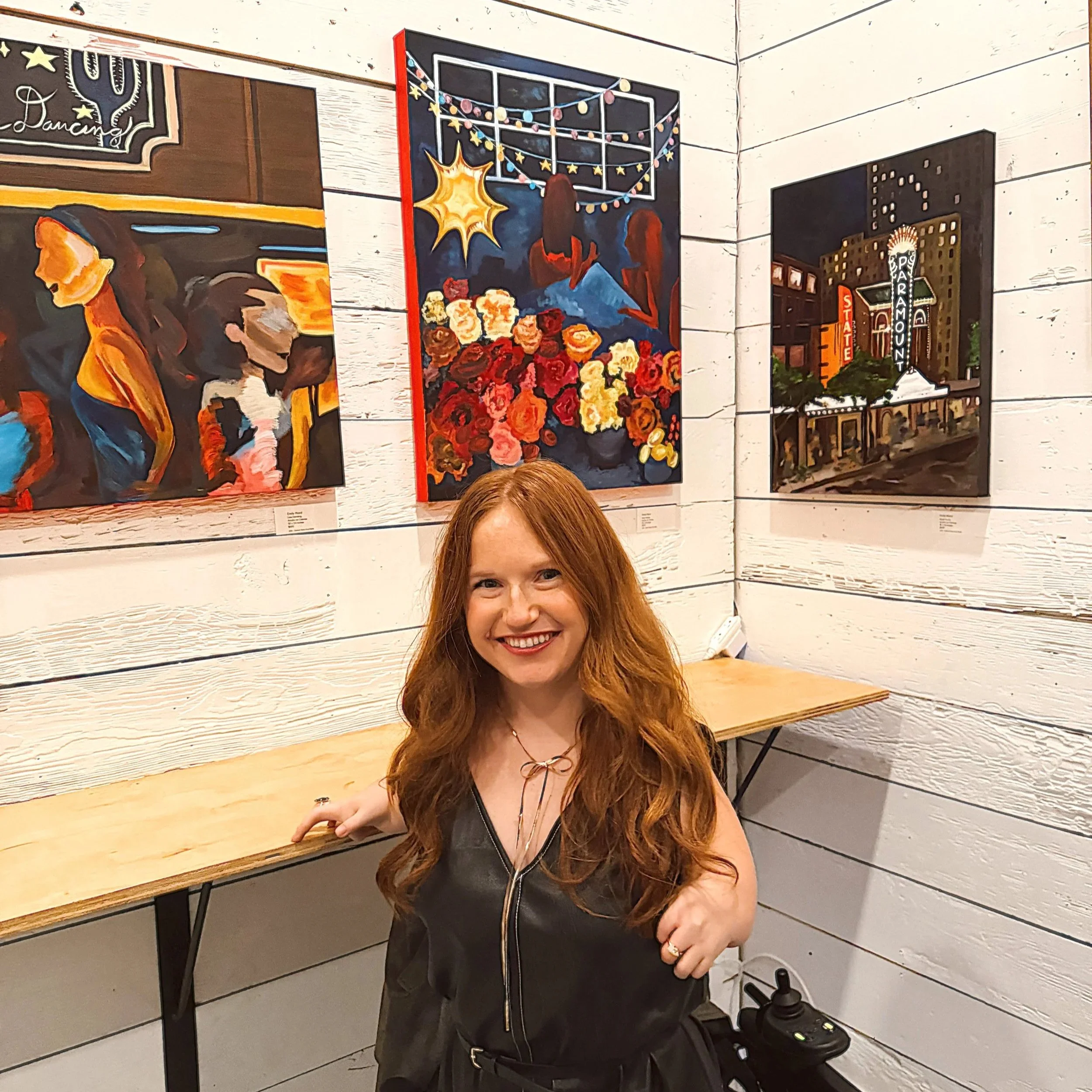 Woman with long red hair smiling at an art gallery, surrounded by paintings on the white wooden wall behind her.