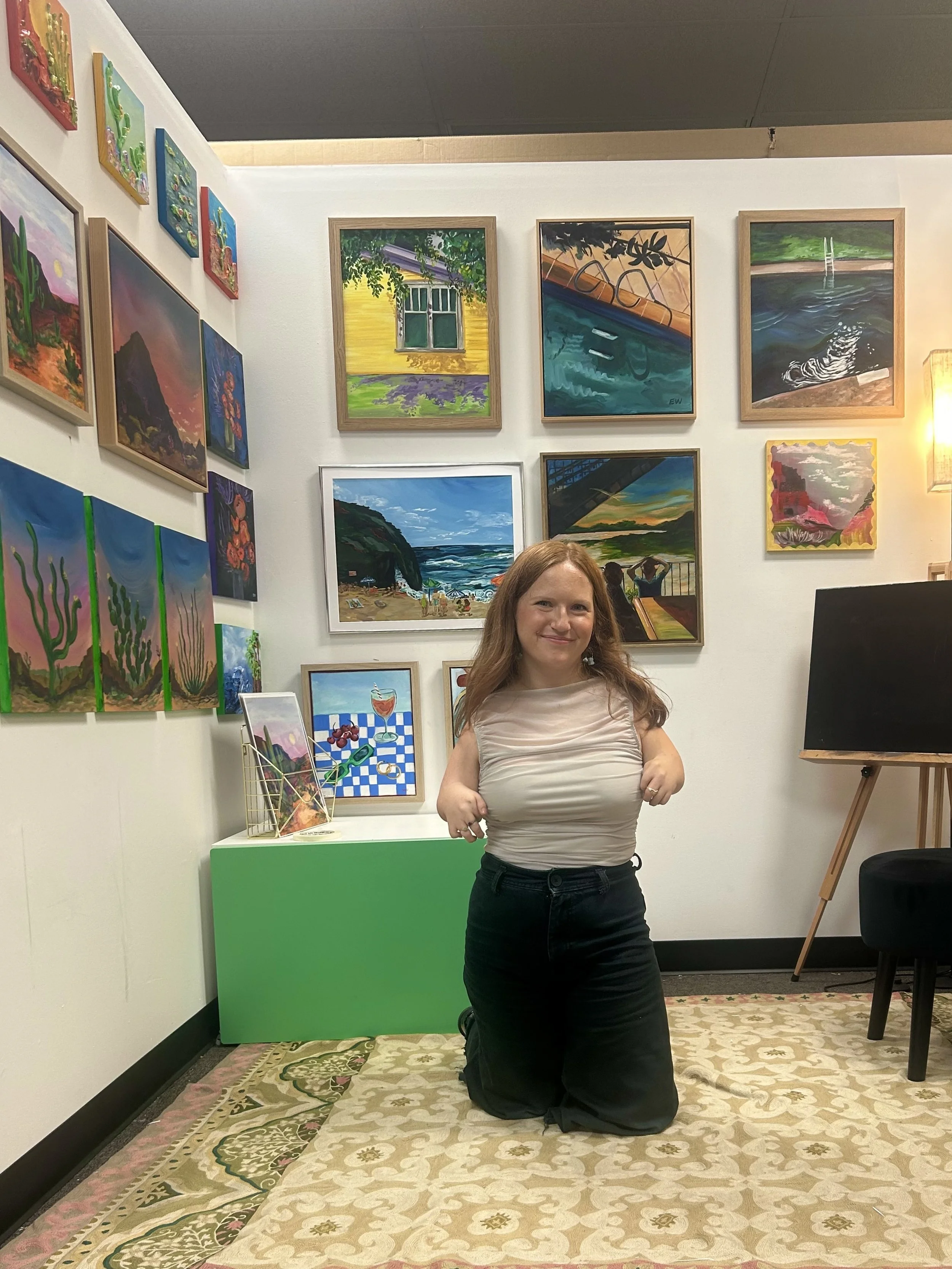 A woman kneeling on a patterned carpet in an art gallery surrounded by colorful paintings on white walls.
