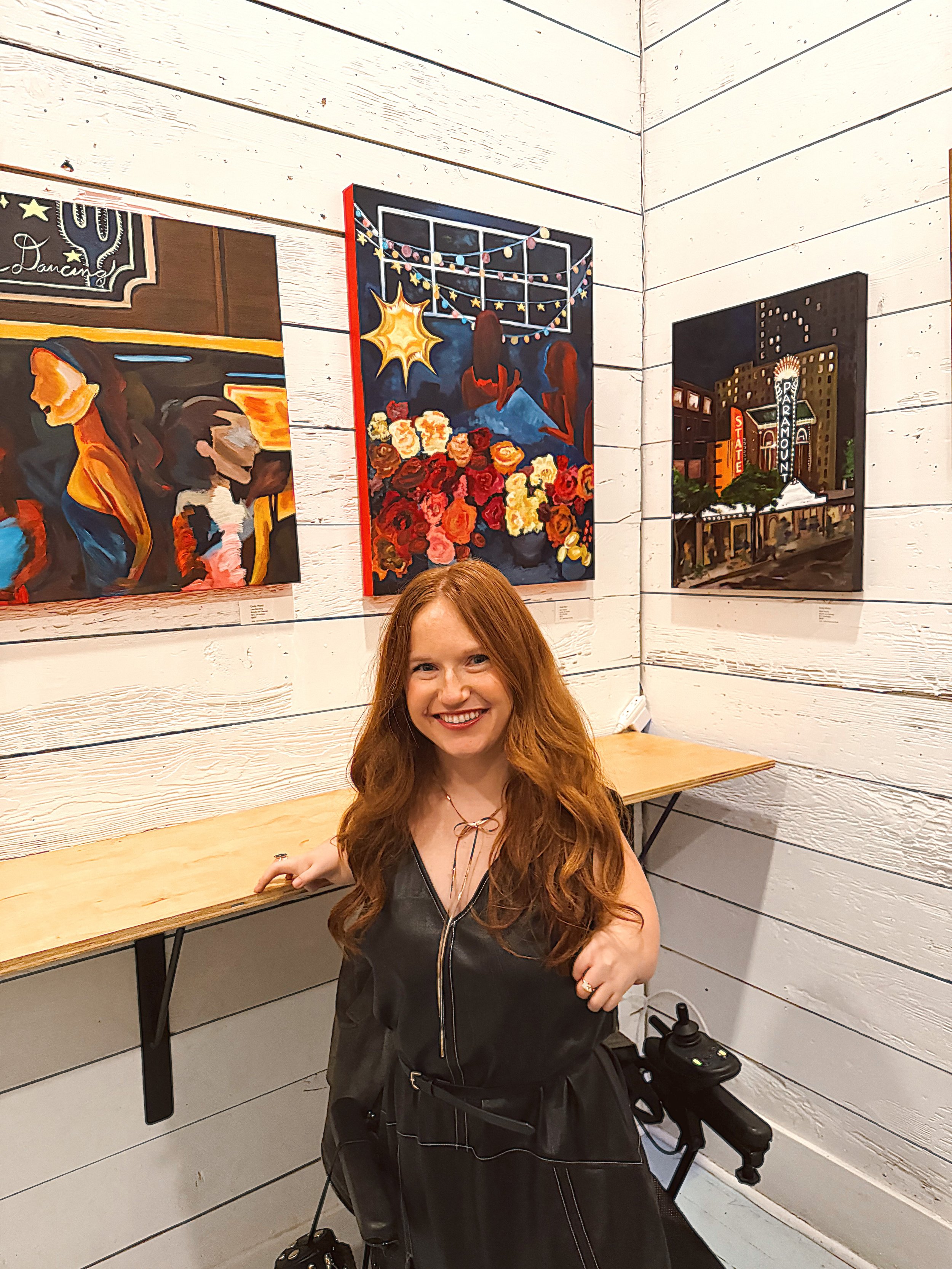 A smiling woman with long red hair sitting in a room with white wooden panel walls, surrounded by colorful paintings hanging on the walls.