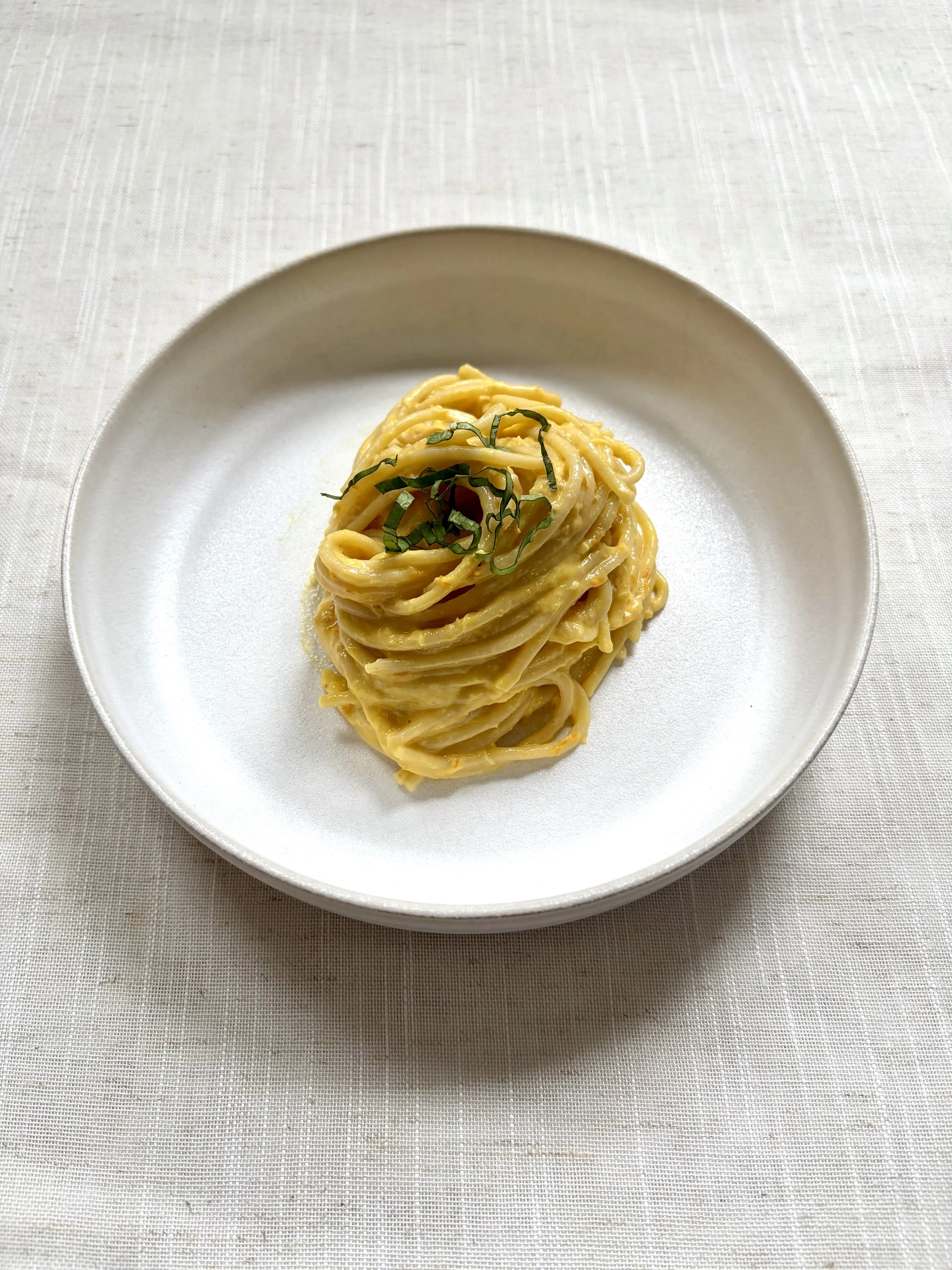 A white plate of spaghetti pasta with a creamy sauce, garnished with basil leaves, on a white tablecloth.