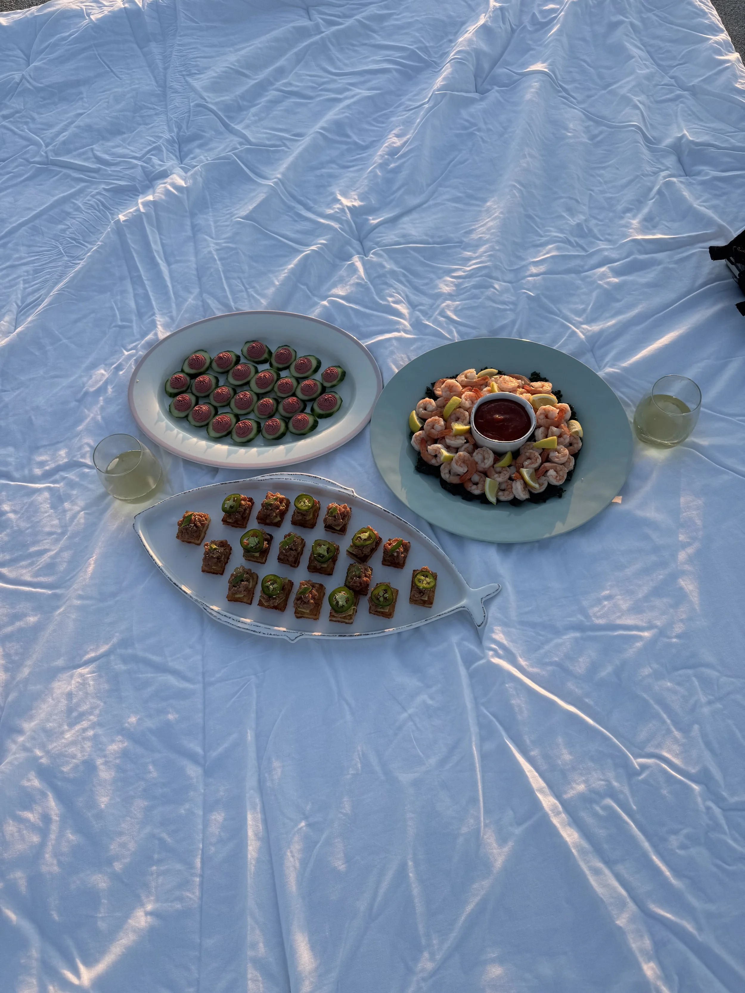 Assorted appetizers on plates, including shrimp with lemon wedges, small green-topped bites, and round pink and green snacks, with drinks on a white tablecloth.