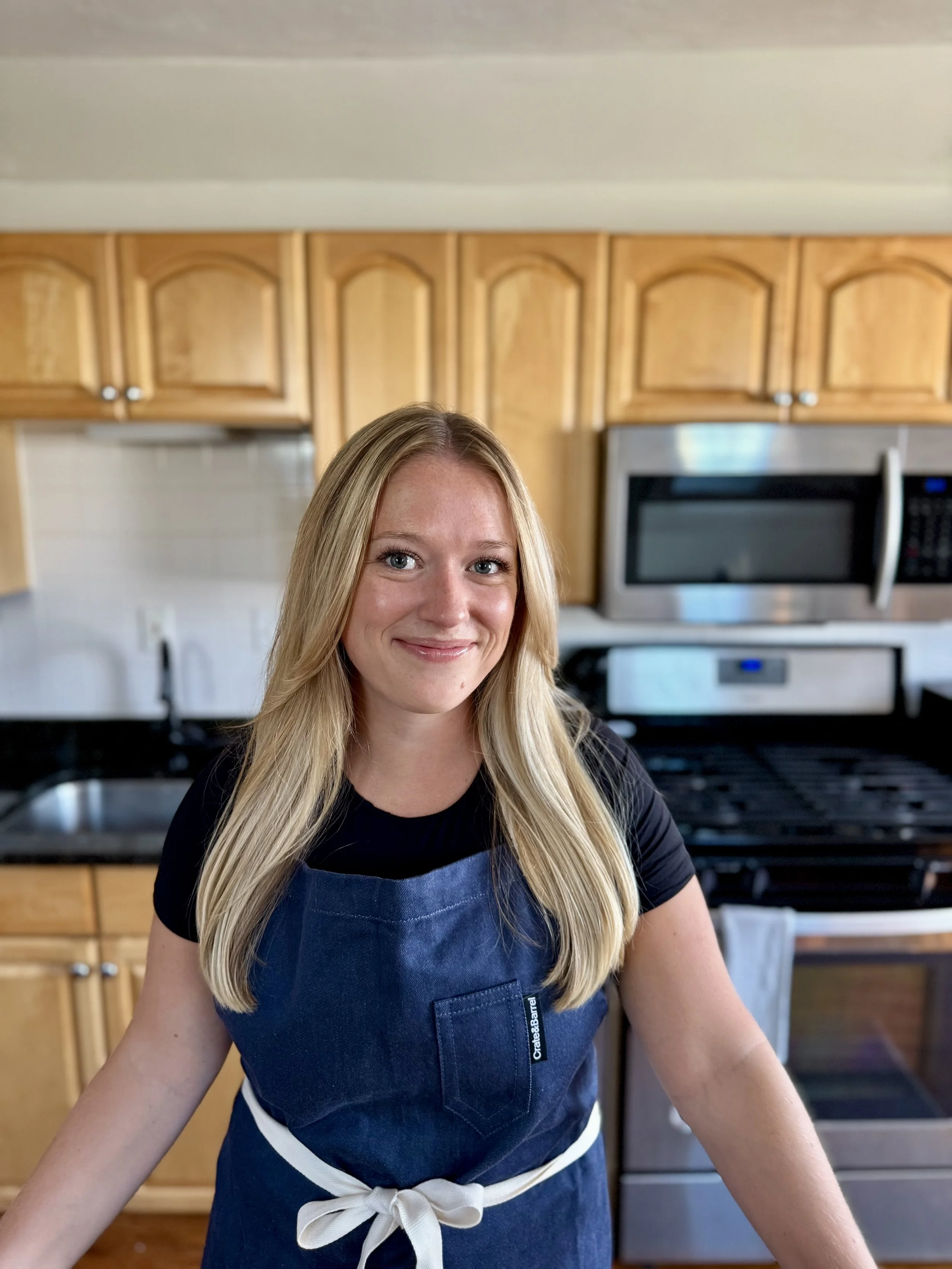 A woman with long blonde hair wearing a black shirt and a blue apron tied at the waist, standing in a kitchen with wooden cabinets and stainless steel appliances.