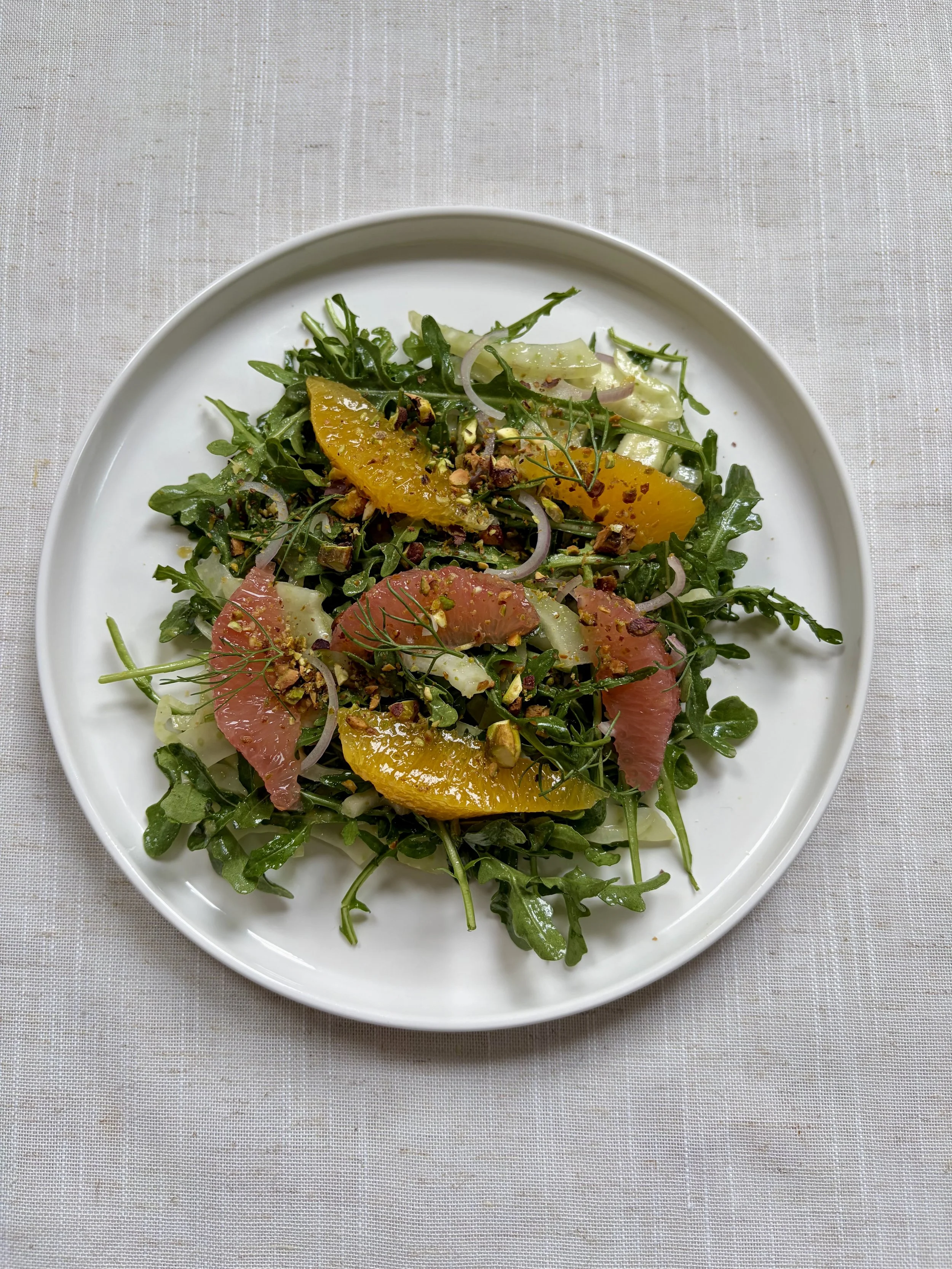 Fresh green arugula salad with slices of grapefruit, orange, and cucumber, garnished with chopped nuts and herbs.