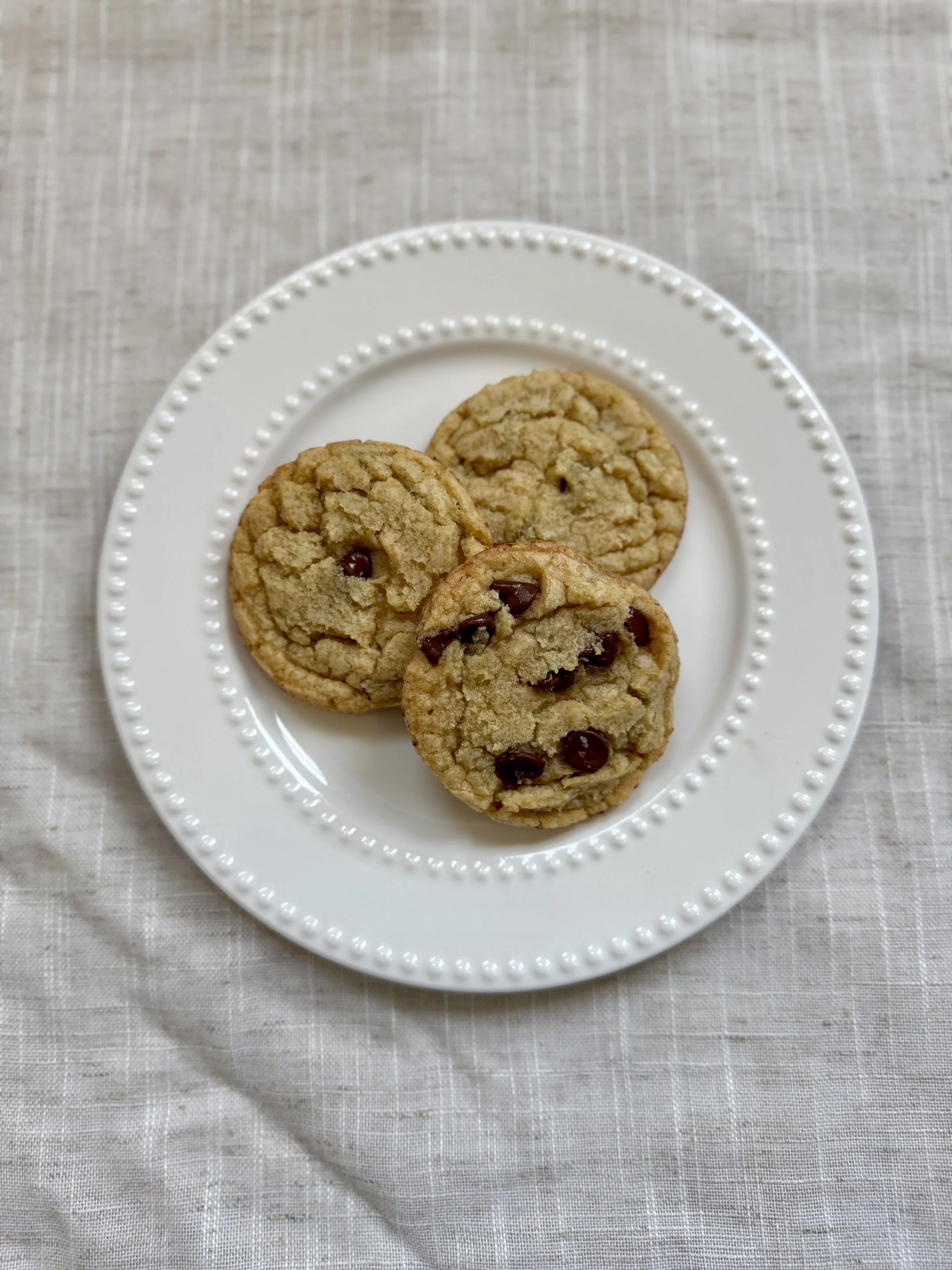 Three chocolate chip cookies on a white decorative plate, placed on a light-colored textured tablecloth.