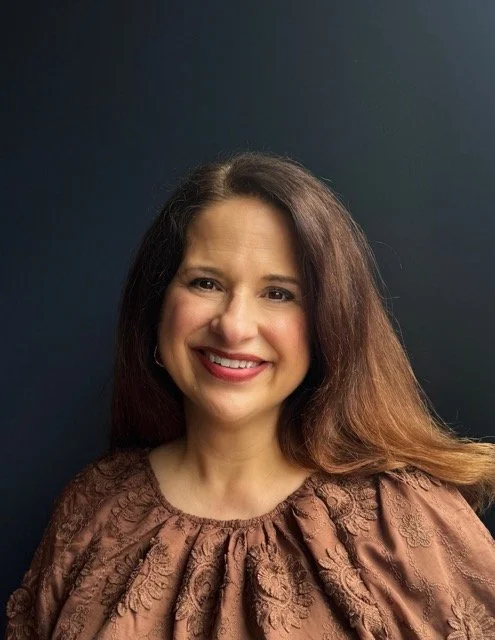 A woman with long brown hair smiling, wearing a brown patterned top, standing against a dark background.