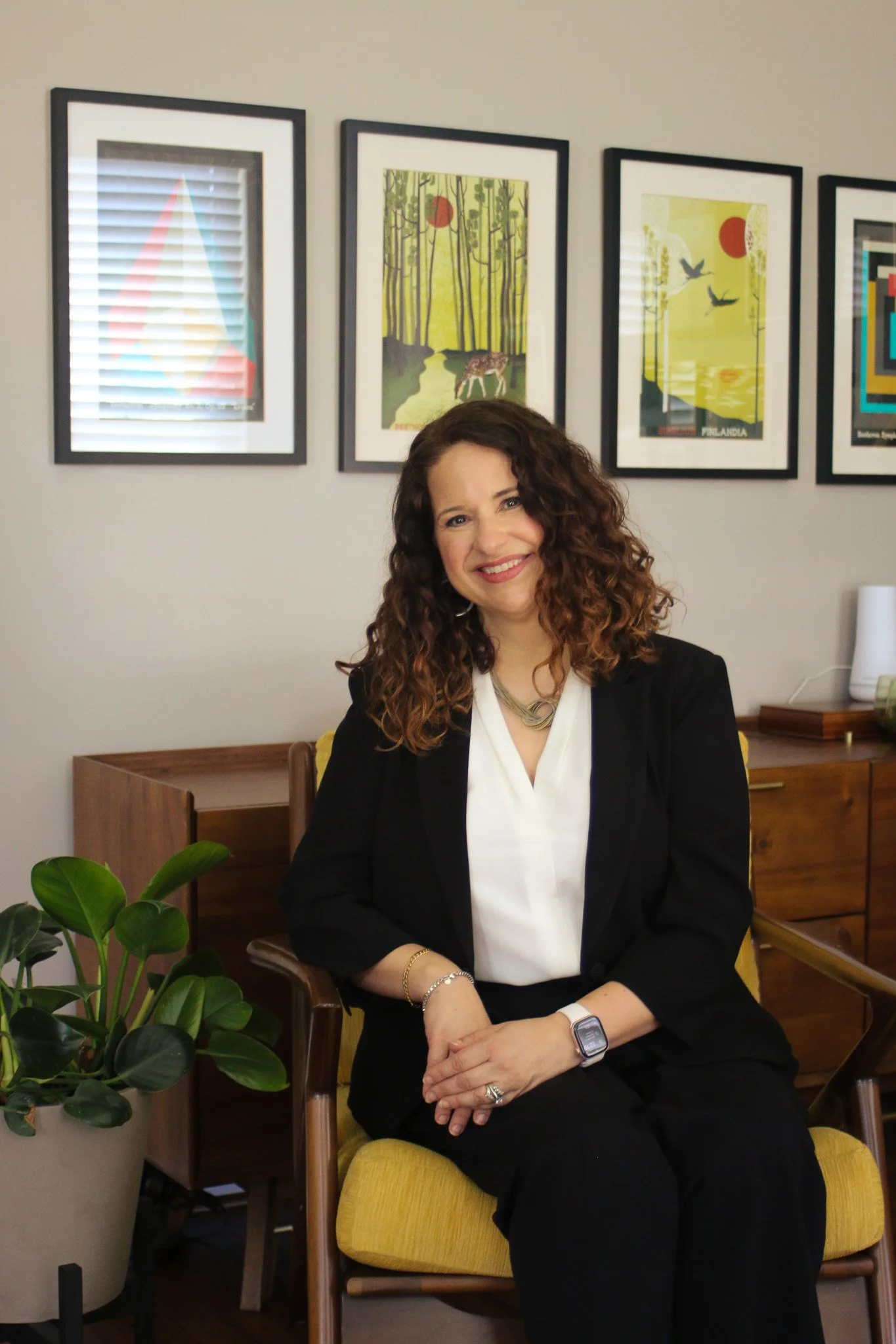 A woman smiling, seated indoors on a yellow chair, with framed artwork on the wall behind her and a houseplant in the foreground.