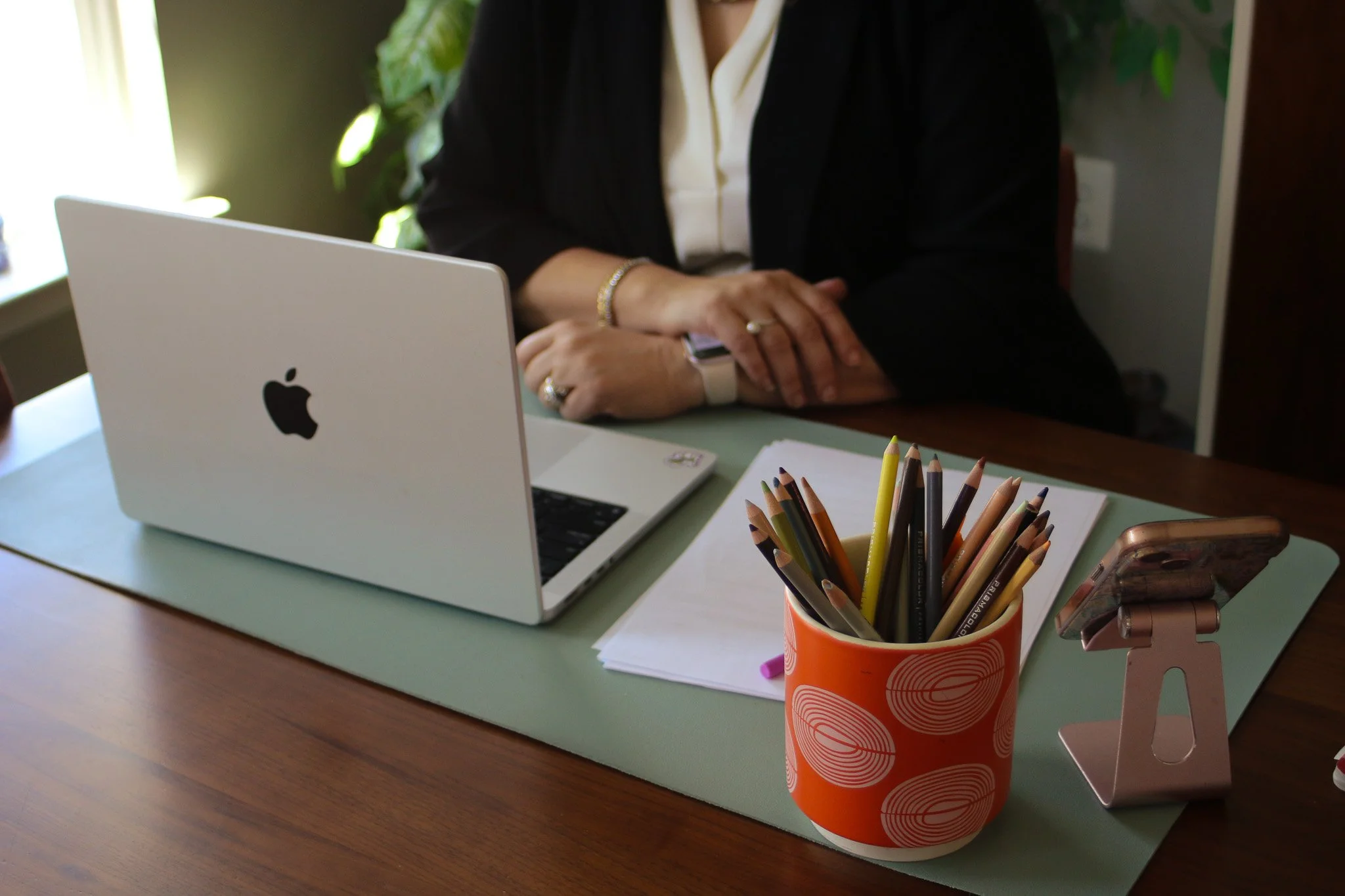A desk with a silver MacBook laptop, a container of colored pencils, a phone, and some sheets of paper. In the background, a person wearing a black blazer and white shirt is seated with their arms crossed, and there are green plants on the window ledge.