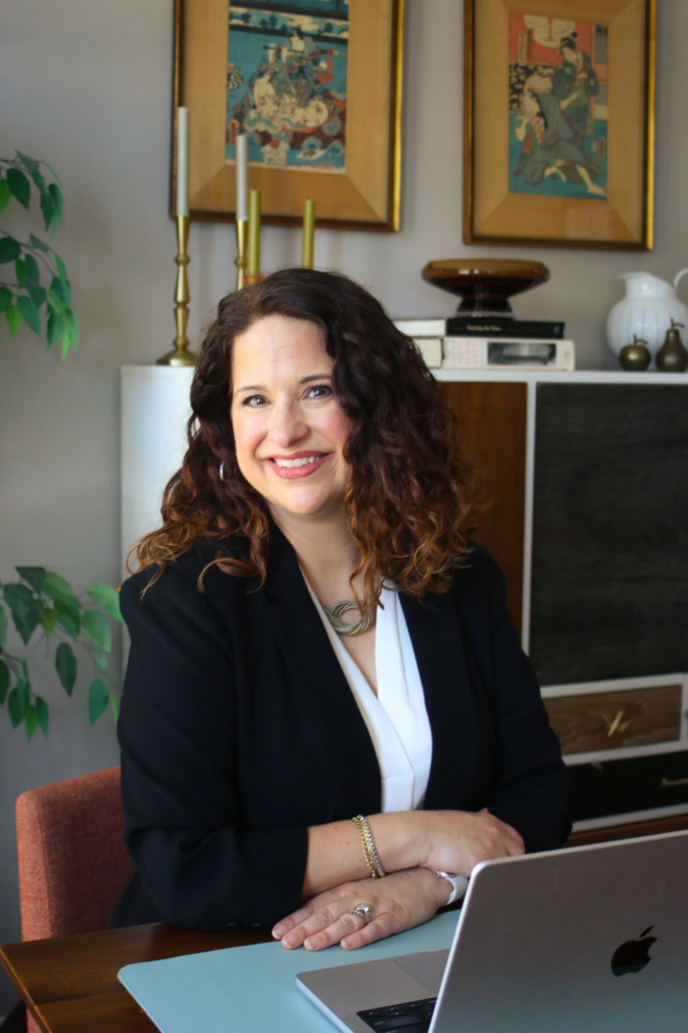 A woman with curly brown hair smiling, sitting at a wooden desk with a silver laptop in front of her. She is wearing a black blazer, white blouse, and a gold necklace. Behind her are framed Japanese art prints, a white cabinet with books and decorative items, and a tall brass candlestick with unlit white candles. To her left, there is a green leafy plant.