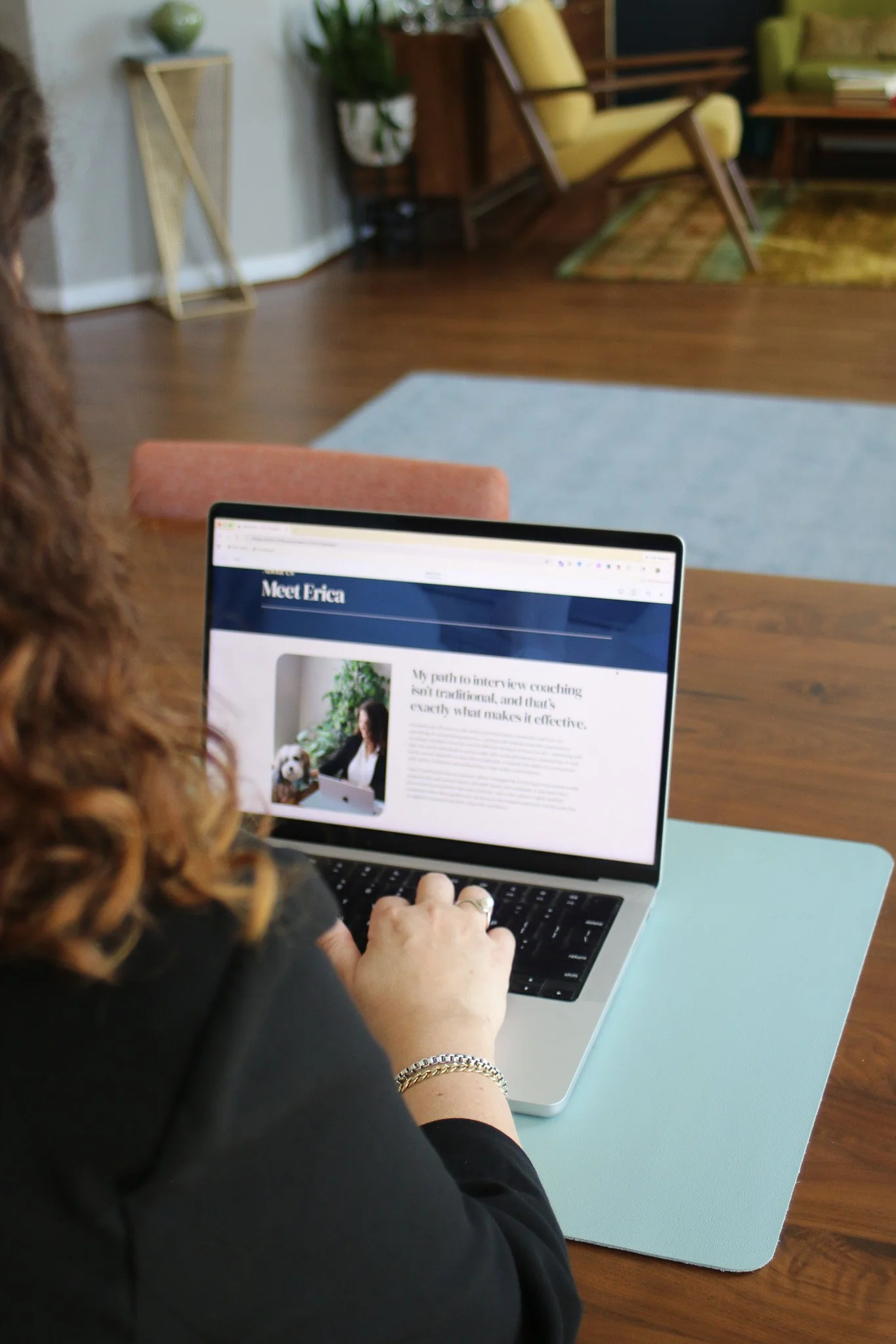 A woman with curly hair using a laptop at a table in a room with wooden floors and mid-century style furniture. The laptop screen displays a website called "Meet Erica" with a picture of a woman and a dog.