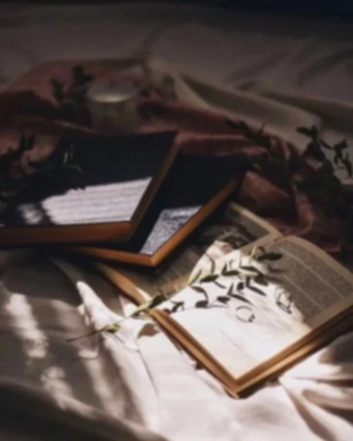 Open books and a tablet placed on a bed with a pink blanket, surrounded by decorative leaves and a lit candle in the background. Marigold's book club.