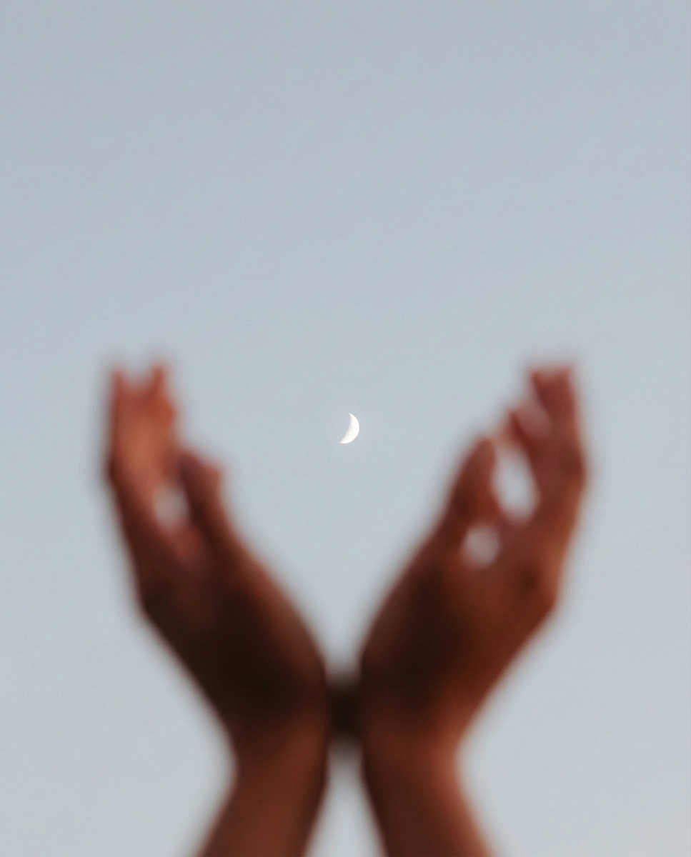 Blurred hands reaching toward a small crescent moon in the sky, evoking stillness, wonder, and quiet reflection.