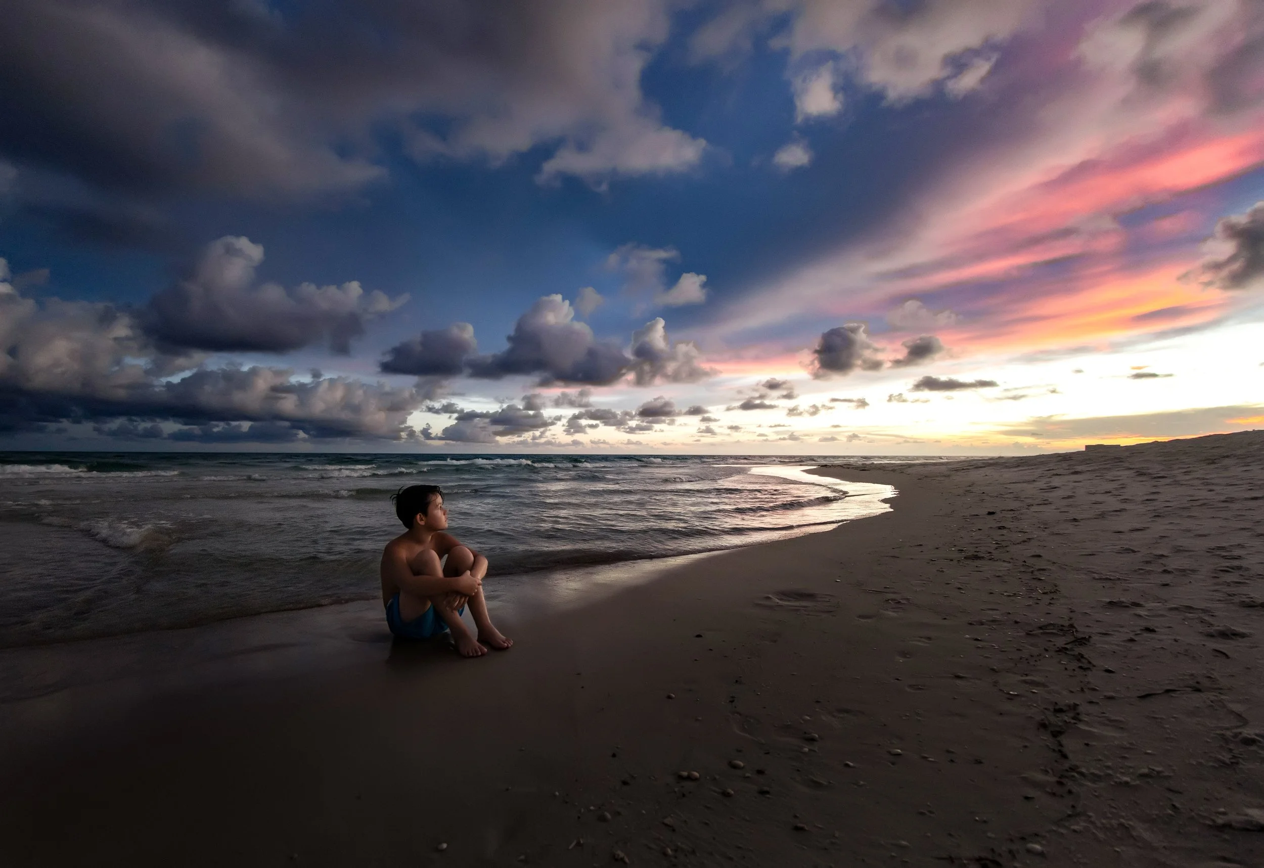 Little boy sitting on he edge of the water on the 30A beach during his sunset family beach session