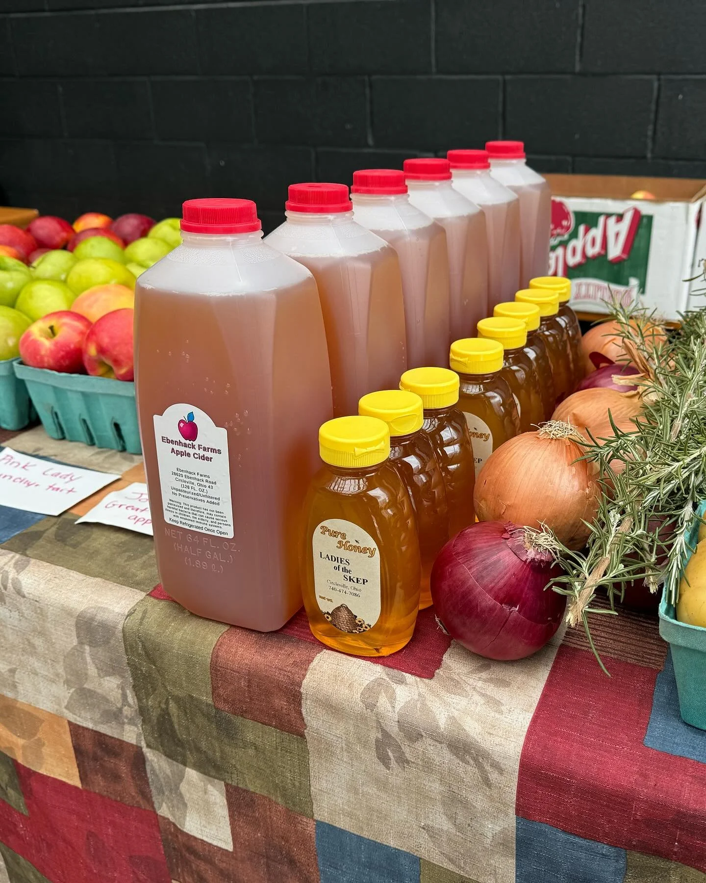 Bottles of apple cider and honey on a table with fresh onions and apples in a market setting.