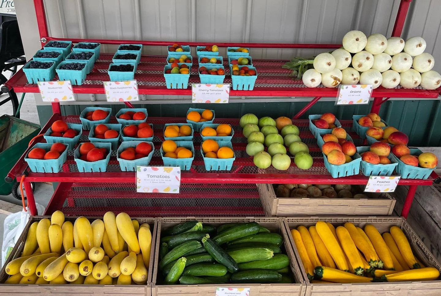 Display of fresh berries, tomatoes, onions, peaches, and zucchinis at a market stand.