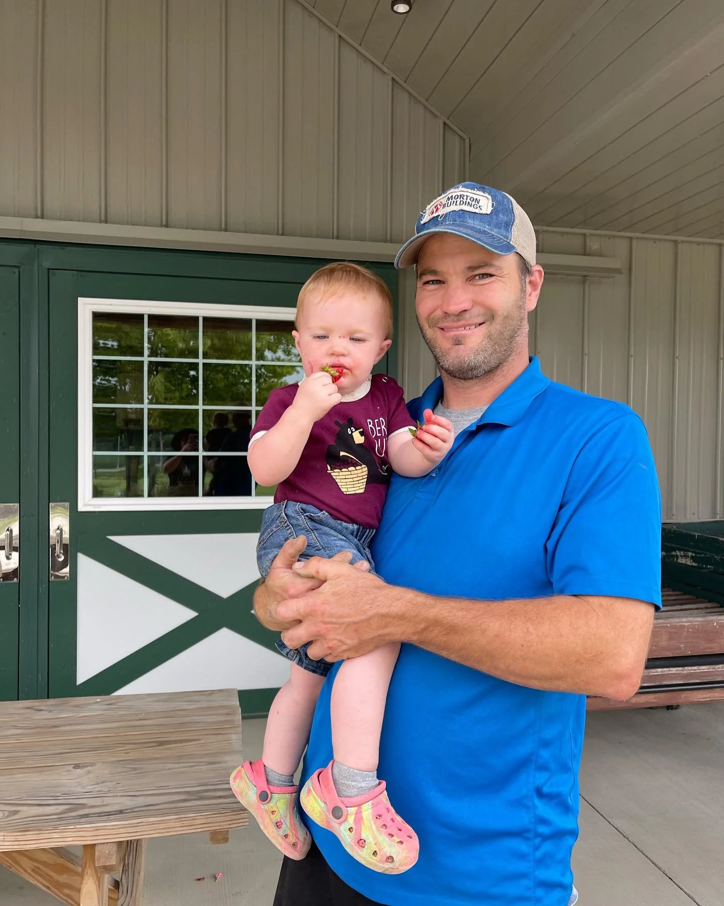 A man wearing a blue shirt and a blue cap holding a young child. The girl is eating a piece of food. They are outdoors in front of a green door with a window, and there is a wooden bench nearby.