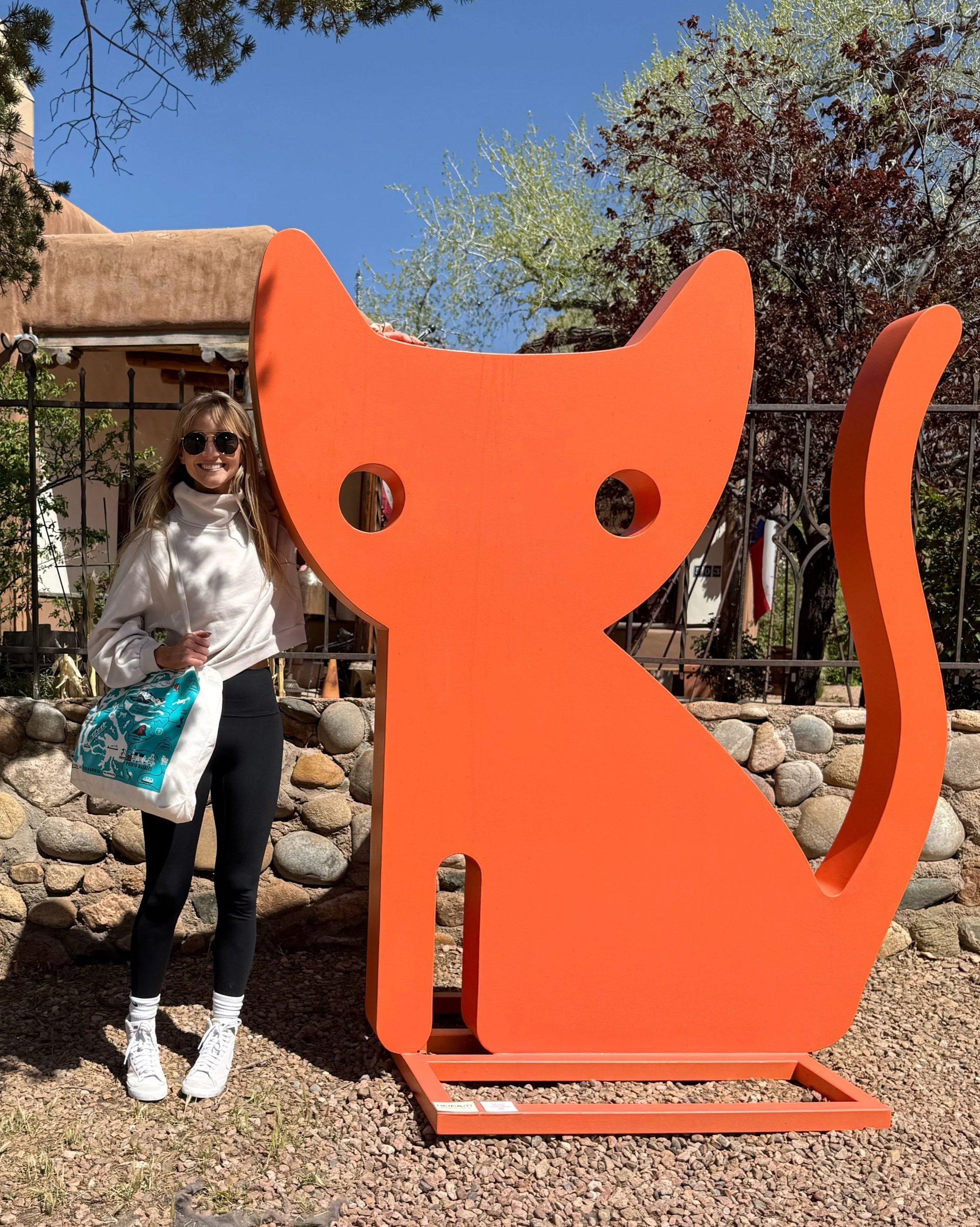 A woman standing next to a large orange cat-shaped sculpture outdoors on a sunny day, with trees and a stone wall in the background.