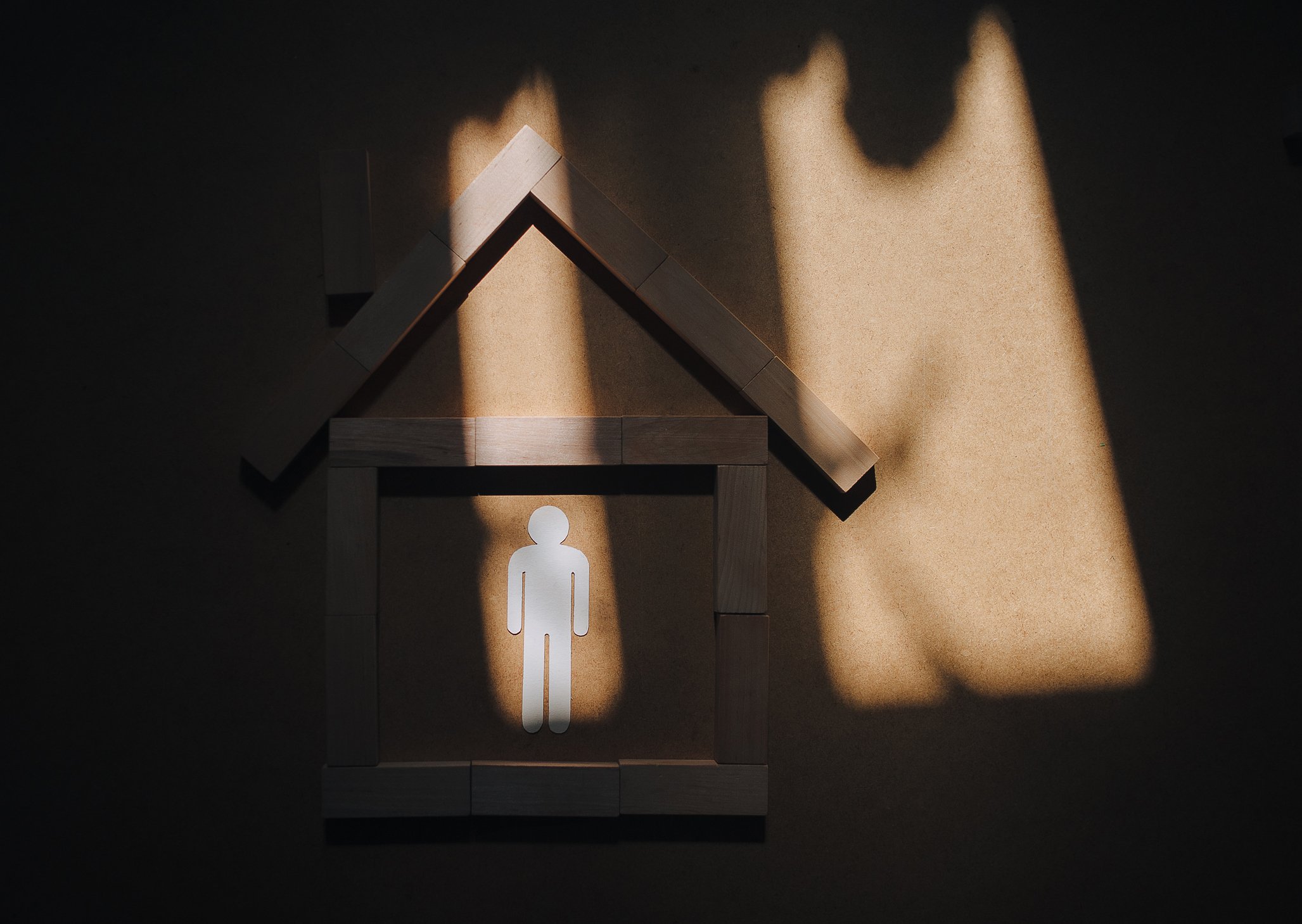 A small house made of wooden blocks with a white cutout of a person inside. Shadows of a roof and window are cast on the wall.
