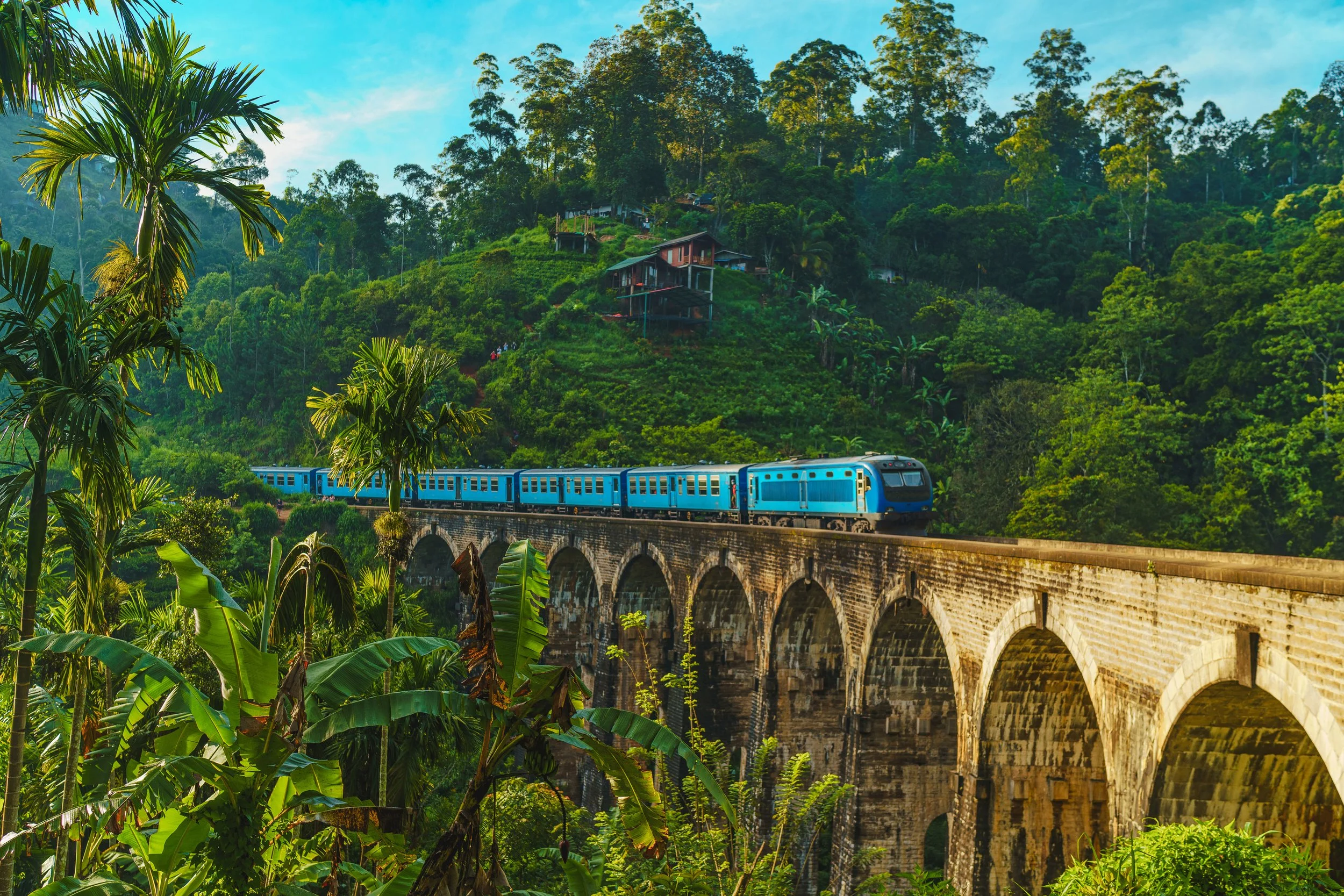 A blue train traveling across a historic stone bridge in a lush, green, tropical landscape with trees and hillside homes.