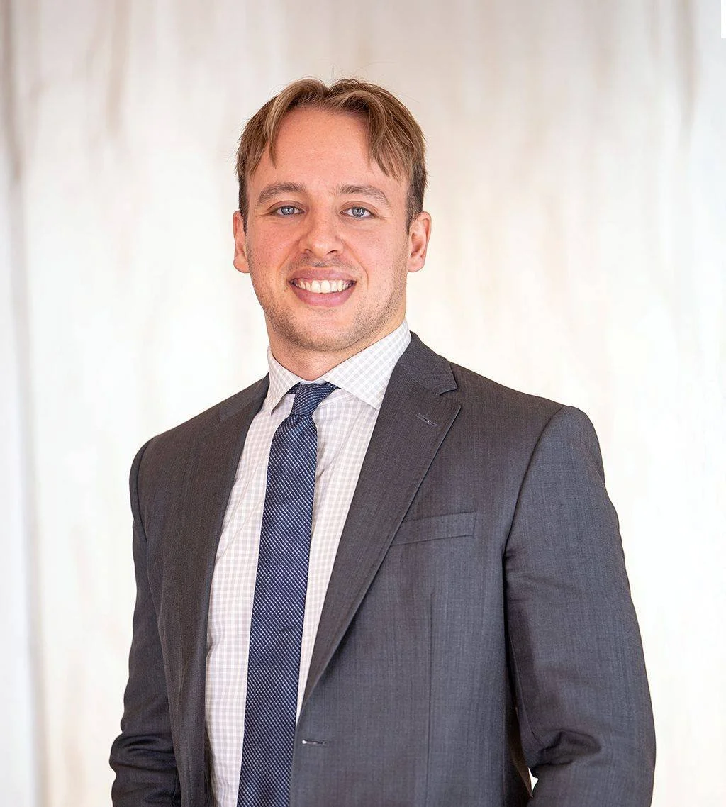 A young man in a dark gray suit, white checkered shirt, and blue tie, smiling and looking at the camera, standing in front of a neutral background.
