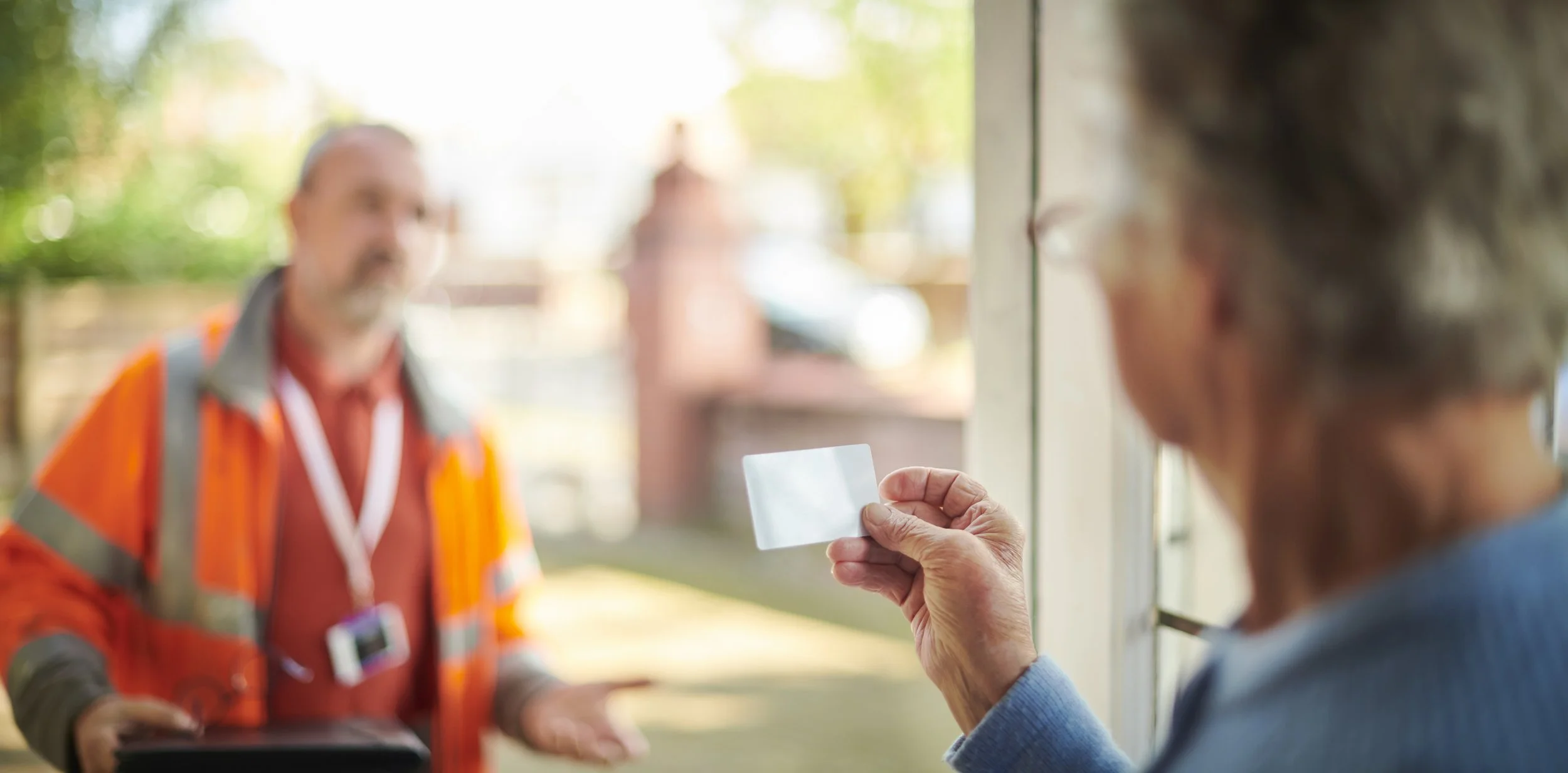 An elderly woman holding a small card or photo, with a man in a bright orange safety vest and orange shirt, blurred in the background outside.