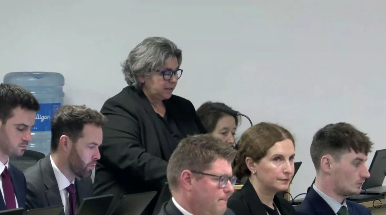A group of people in a meeting room, some working on laptops, with a woman standing and speaking into a microphone, and a water cooler in the background.