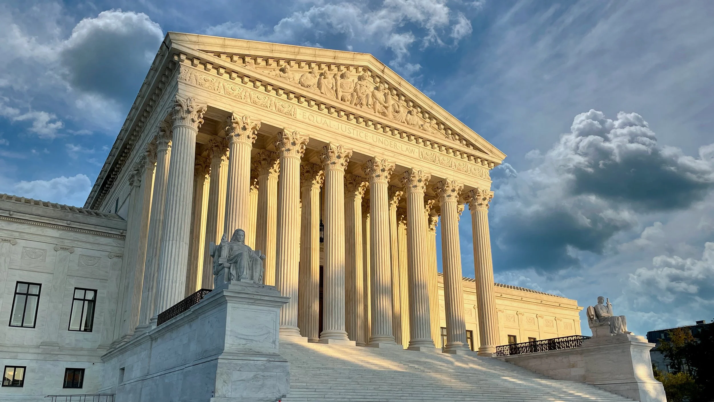 The U.S. Supreme Court building with classical Greek architecture, tall columns, and statues, under a partly cloudy sky, illuminated by sunlight.