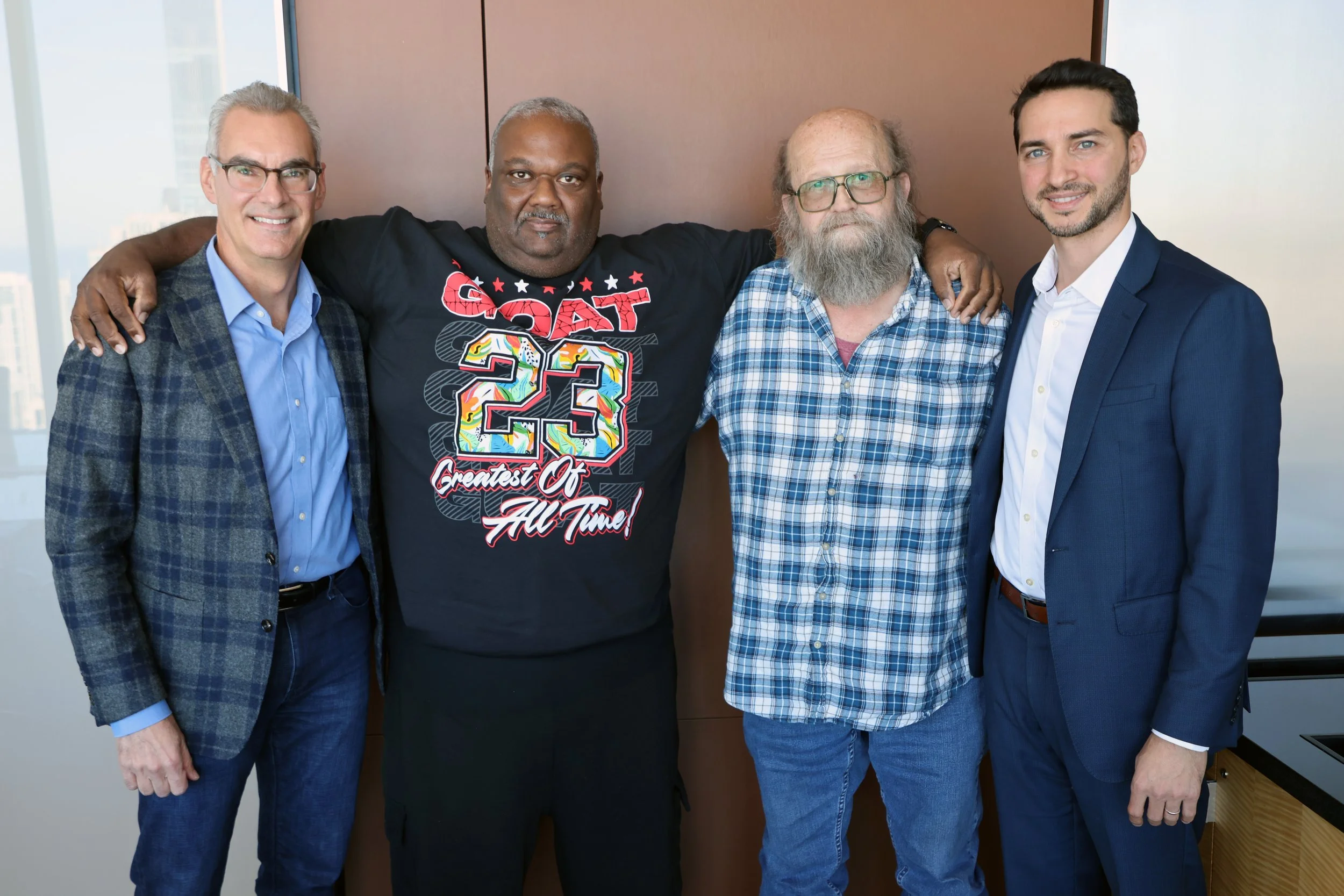 Four men standing together with arms around each other in a meeting room, smiling at the camera.