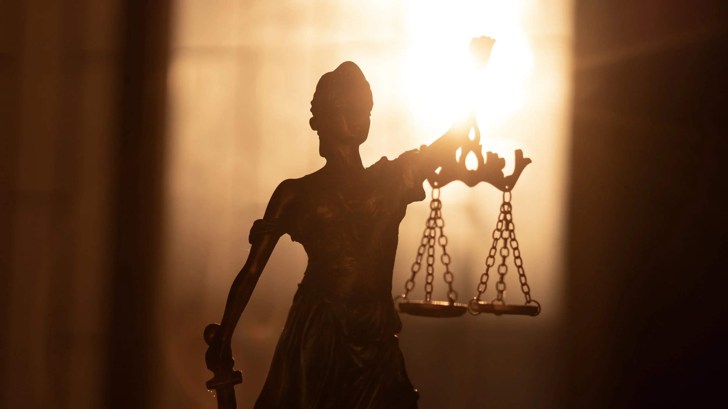 Silhouette of Lady Justice holding a scale, backlit by bright light.