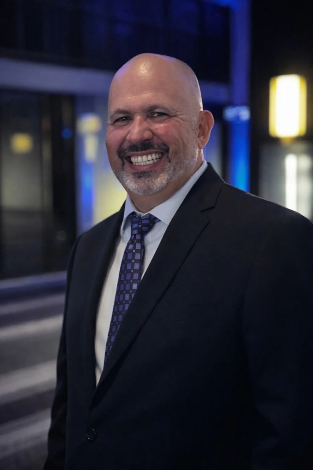 A smiling bald man in a dark suit, white shirt, and purple tie, standing indoors with a blurred modern interior background.