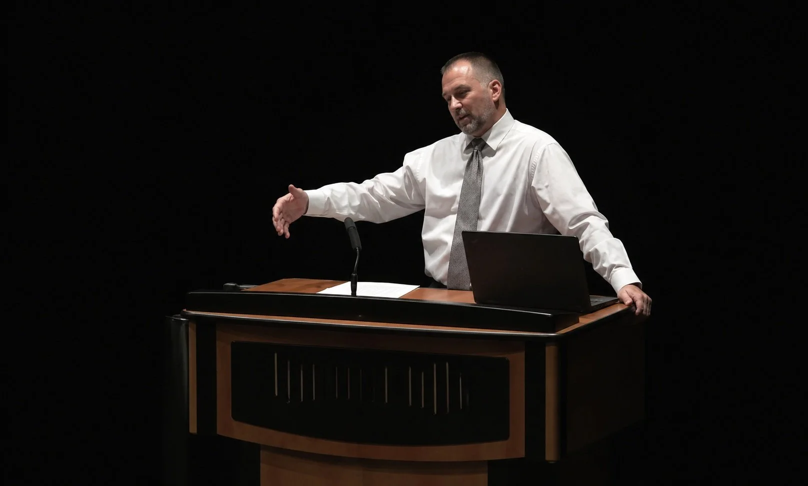 A man in a white shirt with rolled-up sleeves and a gray tie standing behind a wooden podium with a laptop and papers, gesturing with his right hand against a black background.