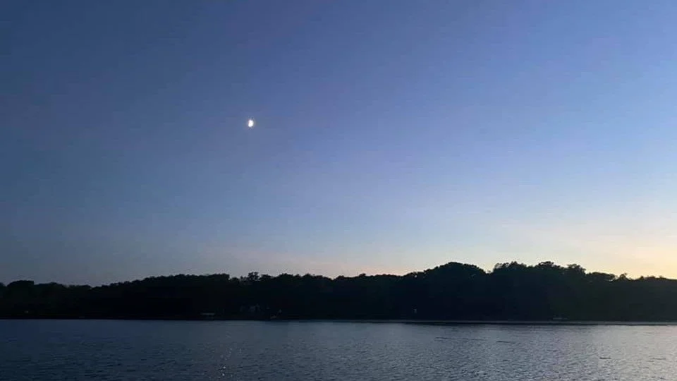 A calm lake at dusk with a dark silhouette of trees on the horizon and a visible moon in the sky.
