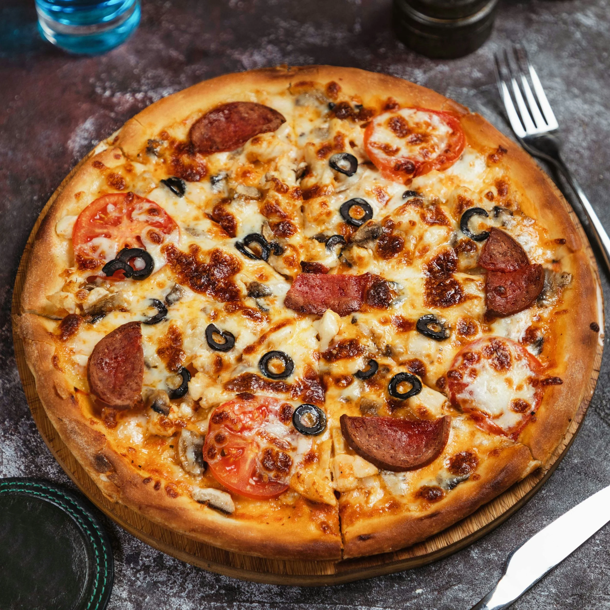 A pizza with pepperoni, tomatoes, black olives, mushrooms, and cheese on a wooden board.