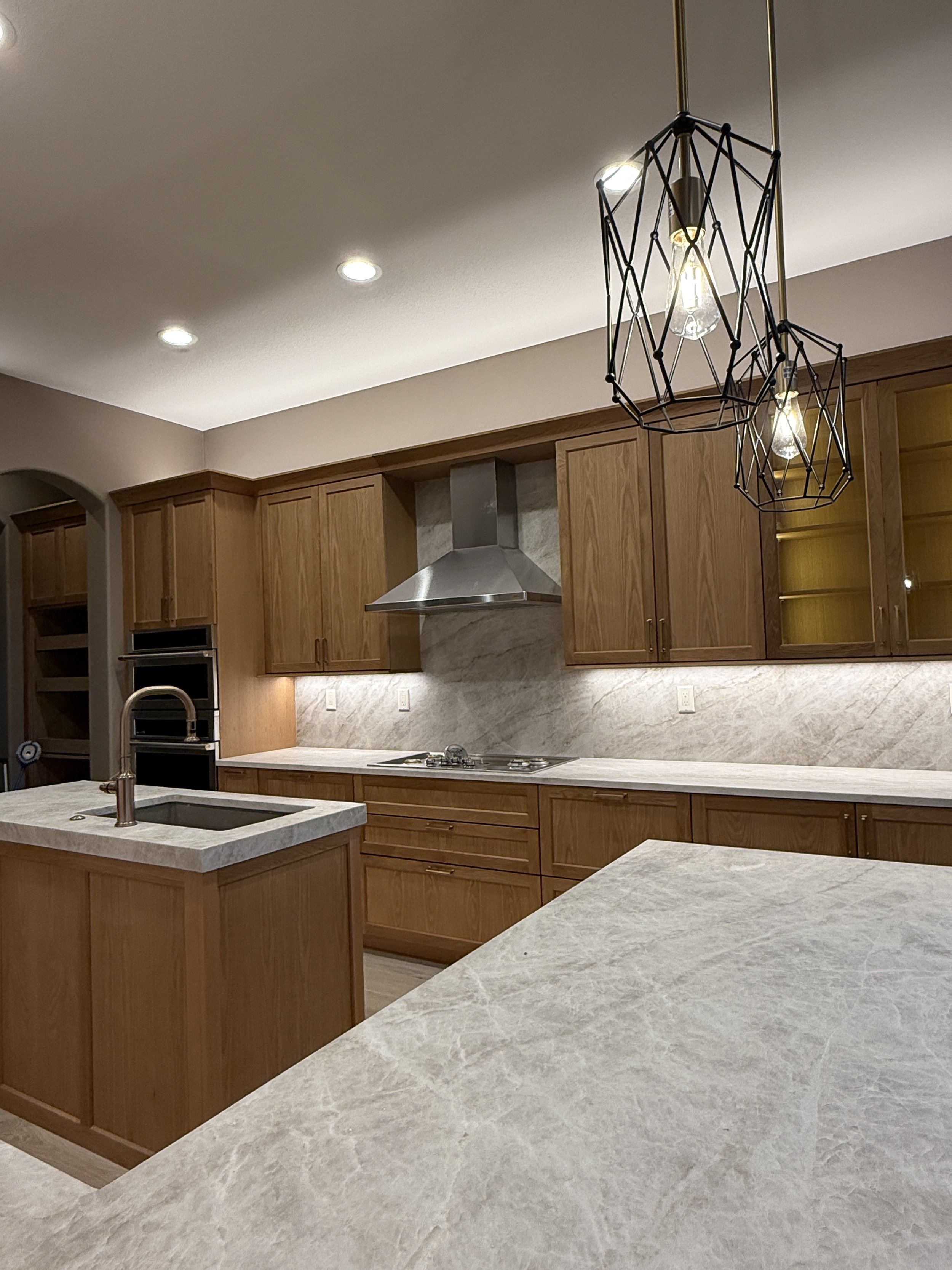 Modern kitchen with wooden cabinets, marble countertops, stainless steel range hood, and black geometric pendant lights.