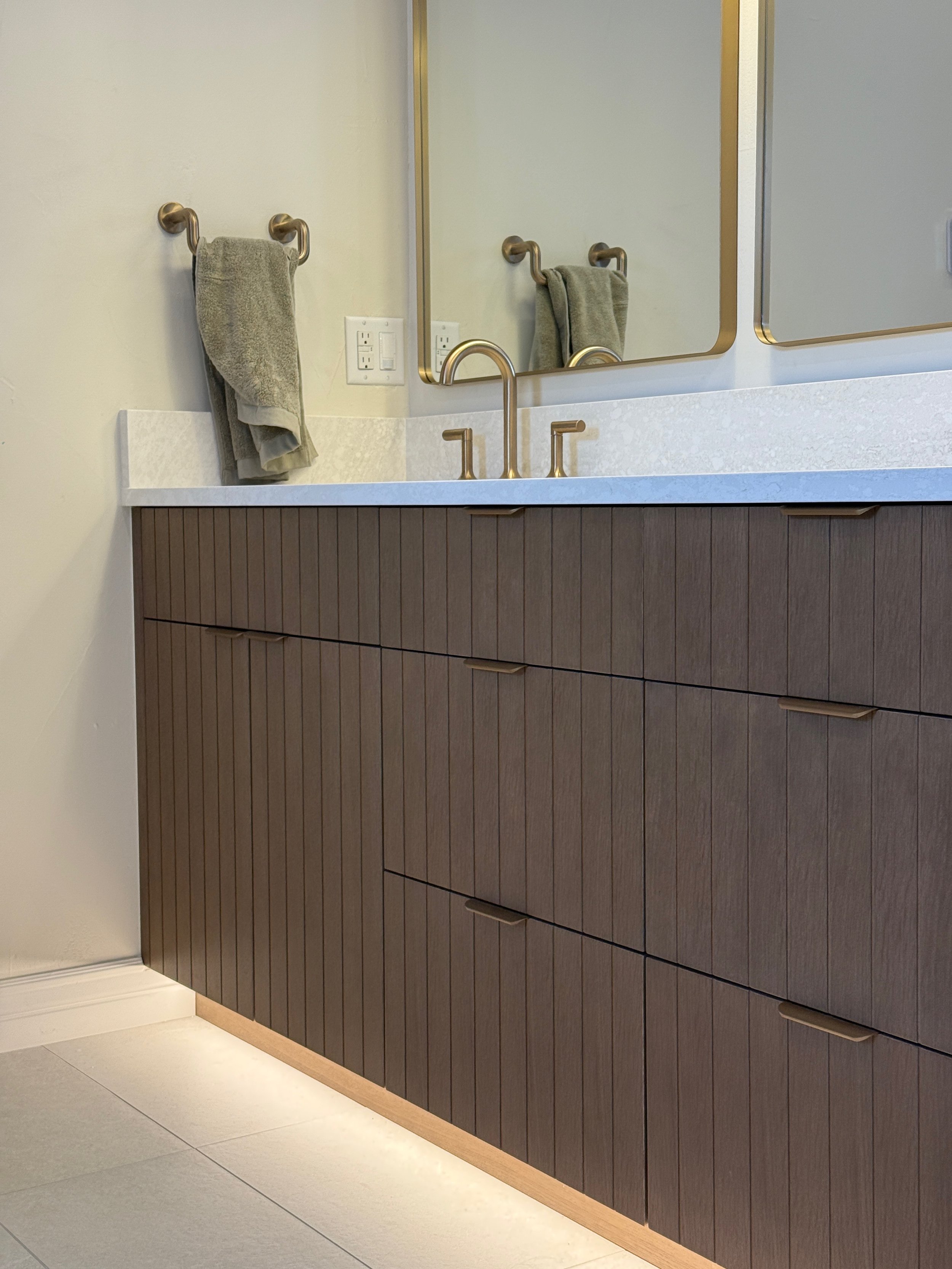 Modern bathroom vanity with brown vertical wood slats, white countertop, two atatable and a wall mirror. A gray towel hangs on a brass towel ring next to the mirror.