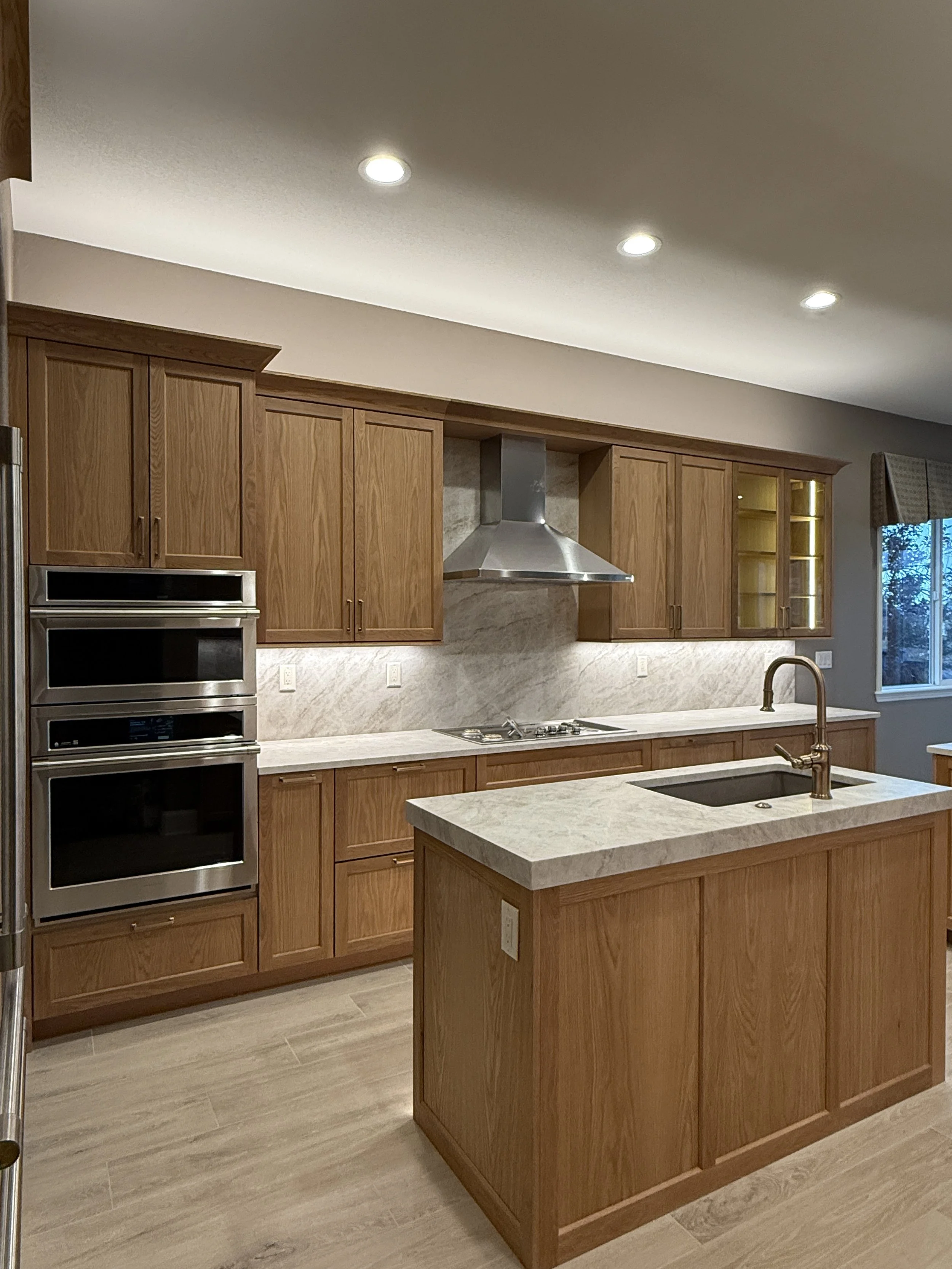 Kitchen with wooden cabinets, a marble island with a sink, a gas cooktop, a stainless steel range hood, and built-in ovens.