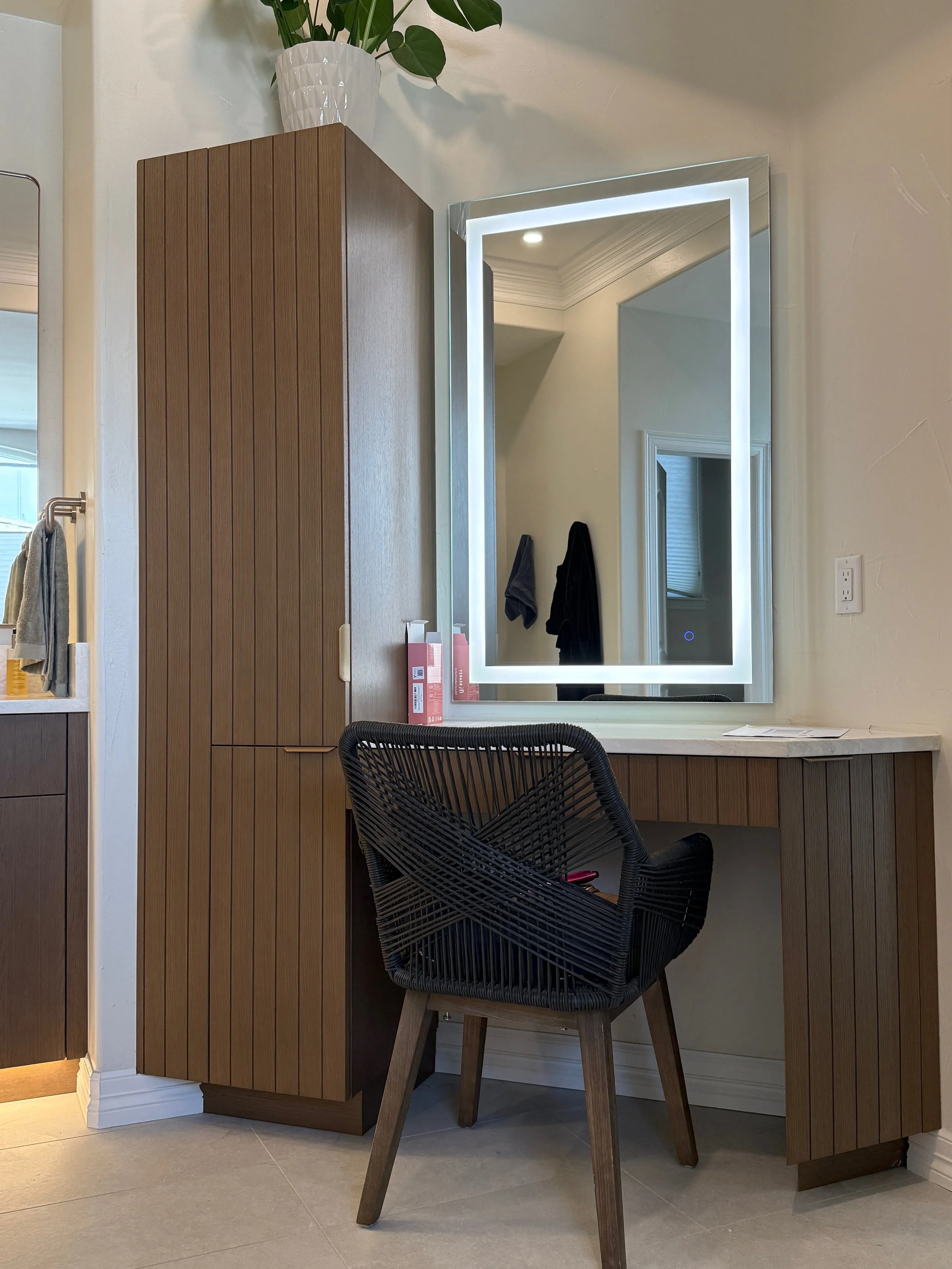 A modern vanity area with a large rectangular mirror with LED lighting, a wooden cabinet and desk with a marble top, a black woven chair, and a mirror on the left wall.
