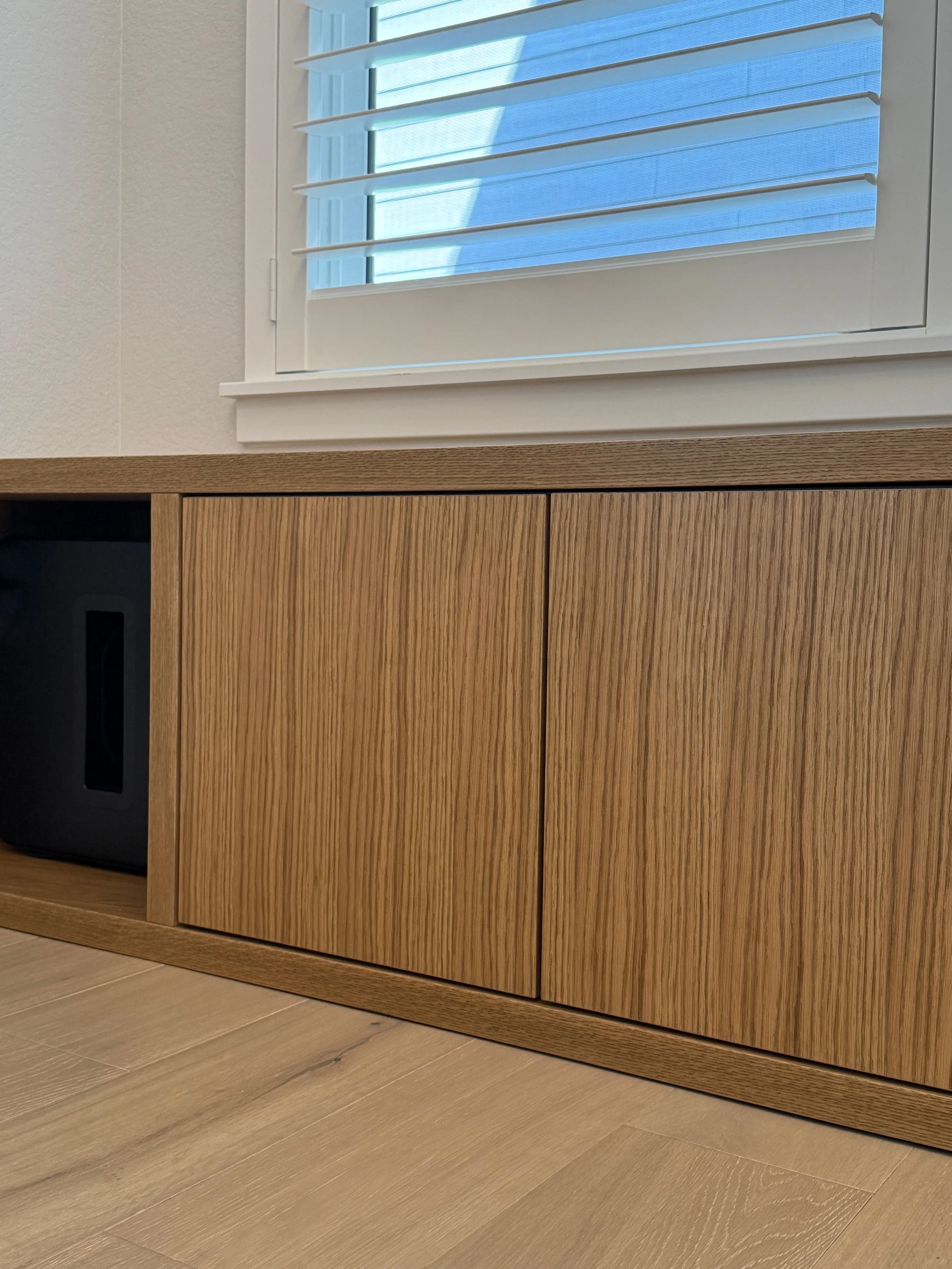 Close-up of a wooden cabinet with closed doors, positioned beneath a window with white blinds, in a room with light-colored wooden flooring and white walls.