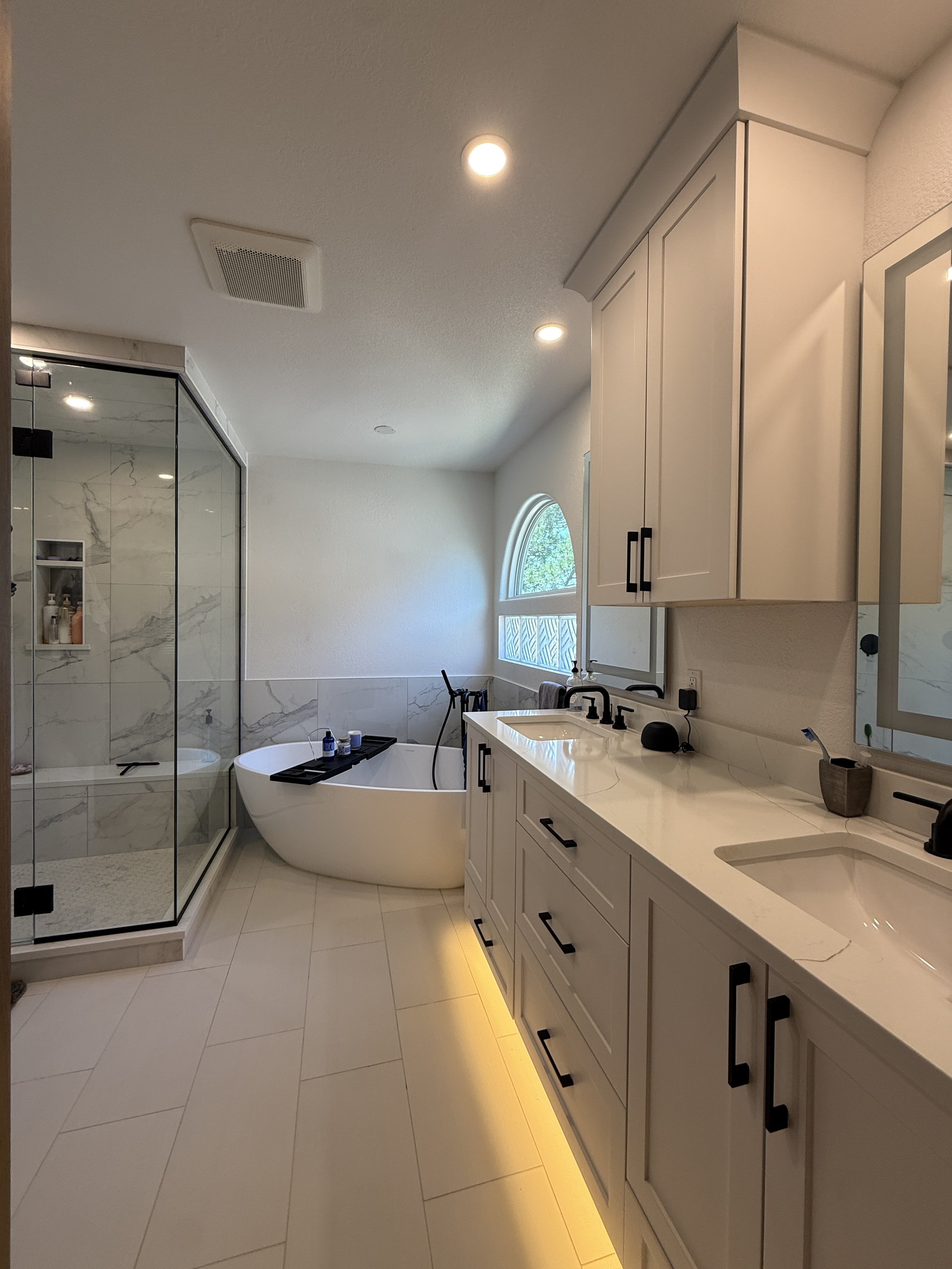 Modern bathroom with white cabinetry, double sinks, a glass-enclosed shower, a bathtub by a window, and under-cabinet lighting.