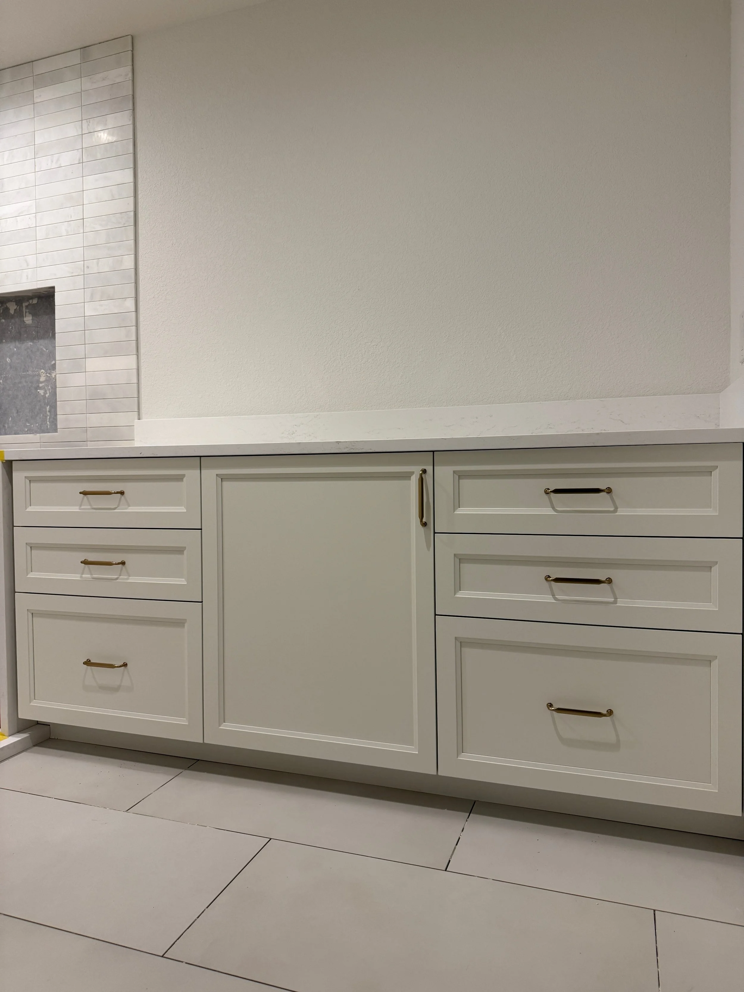 Kitchen cabinet with gold handles and a white countertop, with a wall on top that includes a textured white wall and tiles on the left side.