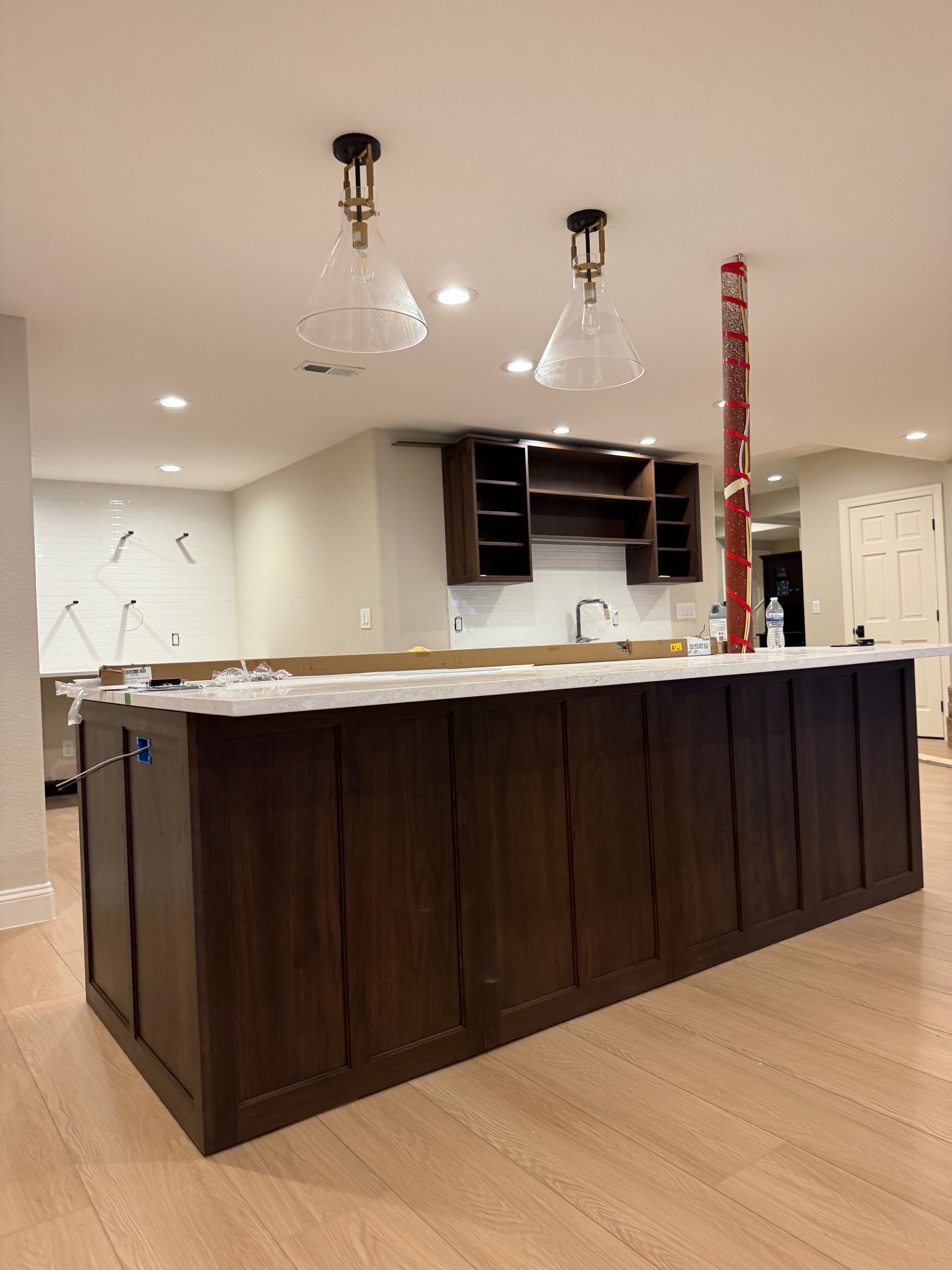 Kitchen island with dark wood cabinets, white marble countertop, and open overhead shelves. Two glass pendant lights hang from the ceiling, and a red decorative column is on the right side. The background shows a white wall with hooks and a clean, mo