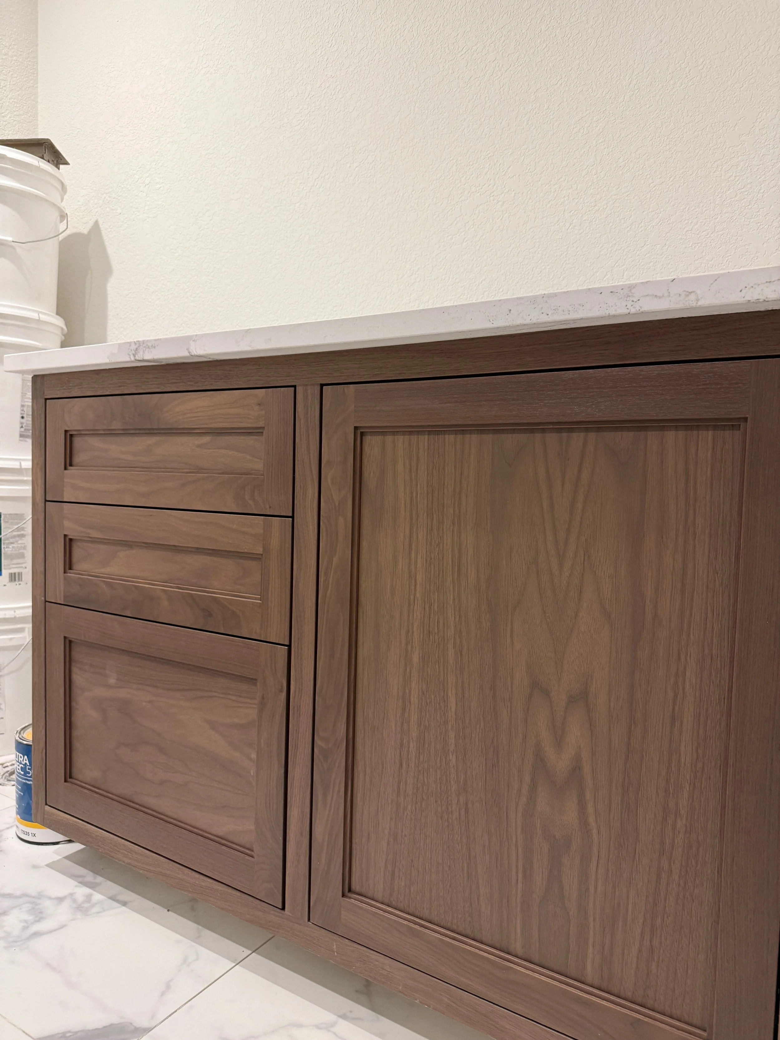 Wooden cabinet with drawers and a door, topped with a white marble countertop, in a room with white walls and marble flooring.