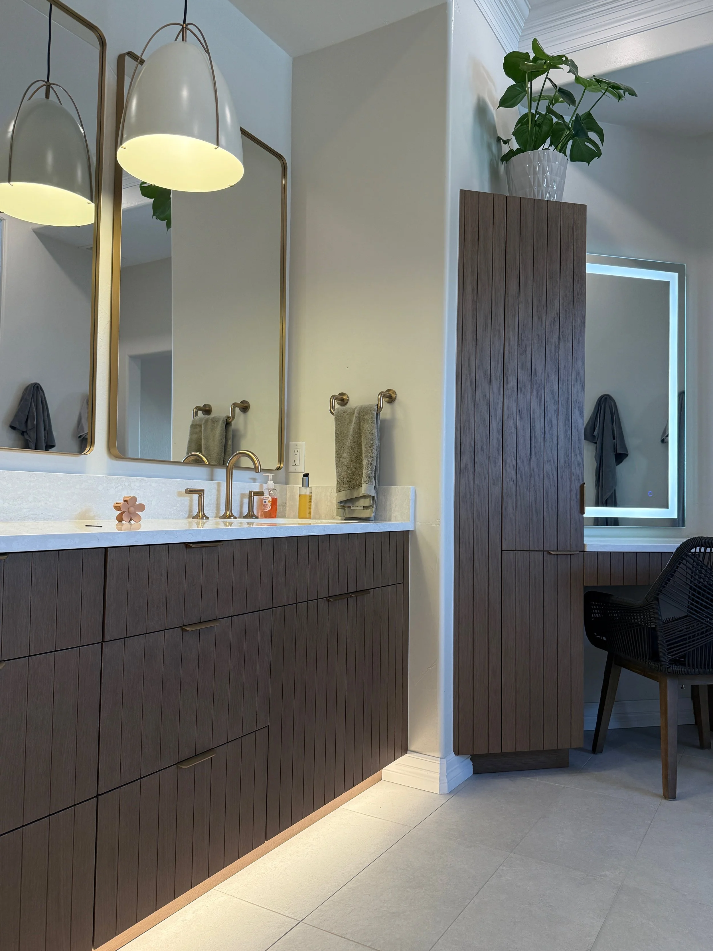 Modern bathroom vanity with wood grain cabinets, a white countertop, and gold fixtures. Two large mirrors with gold frames are above the sink, and pendant lights hang from the ceiling. There are towels on gold towel bars and a potted plant on a tall,