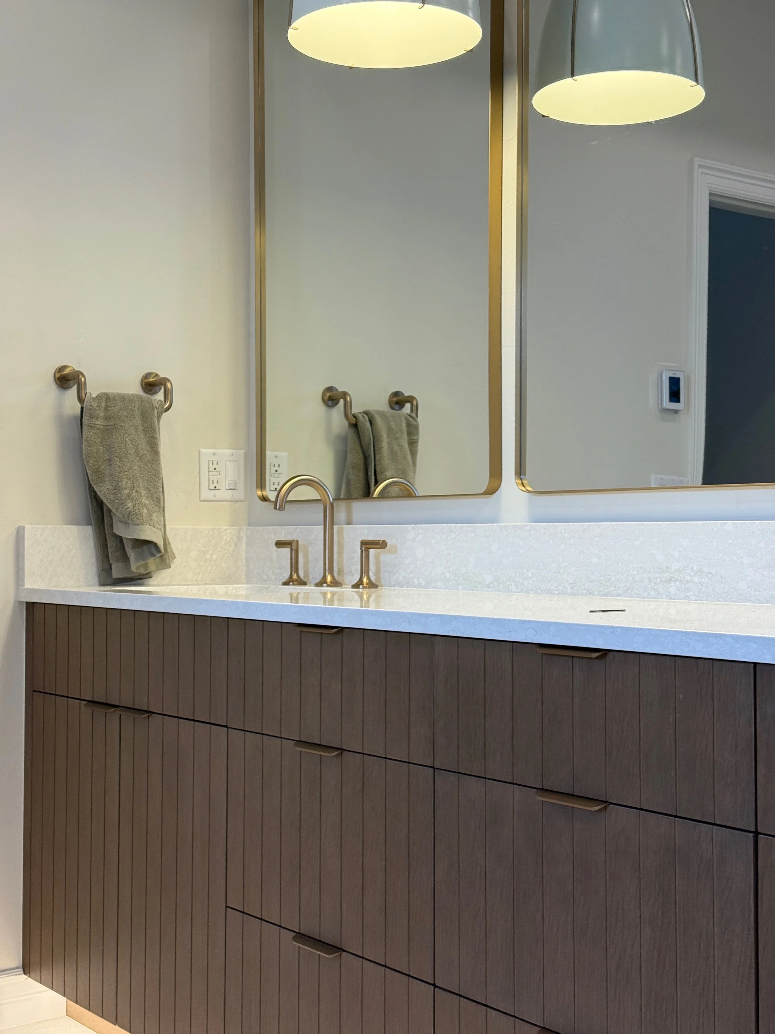 Modern bathroom vanity with a white countertop, brown cabinet drawers, a brass faucet, a large mirror, two pendant lights, and a gray towel hanging on a brass towel bar.