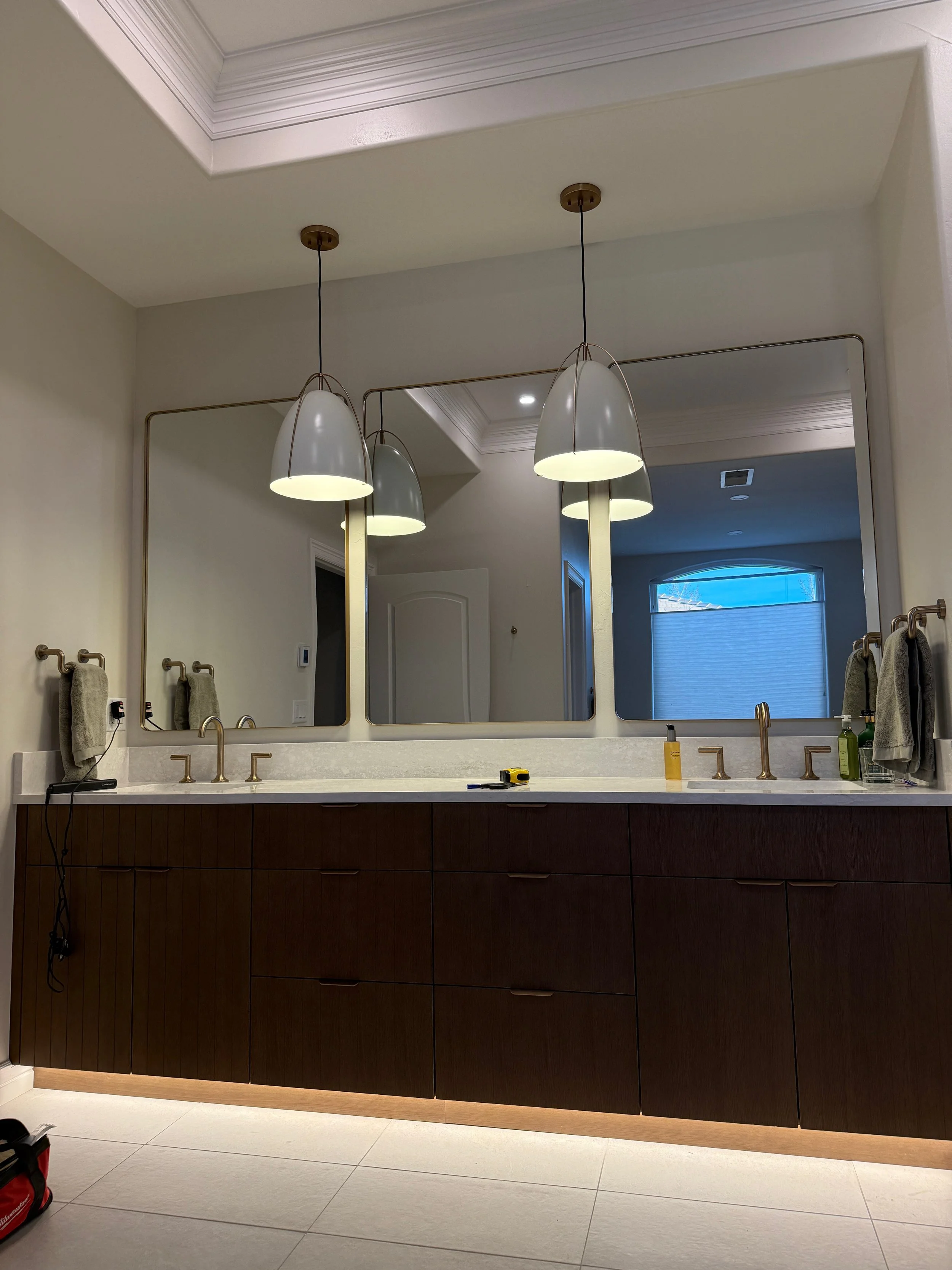 Modern bathroom vanity with two large mirrors, three pendant lights, brass faucets, and wooden cabinetry, with towels and toiletries on the countertop.