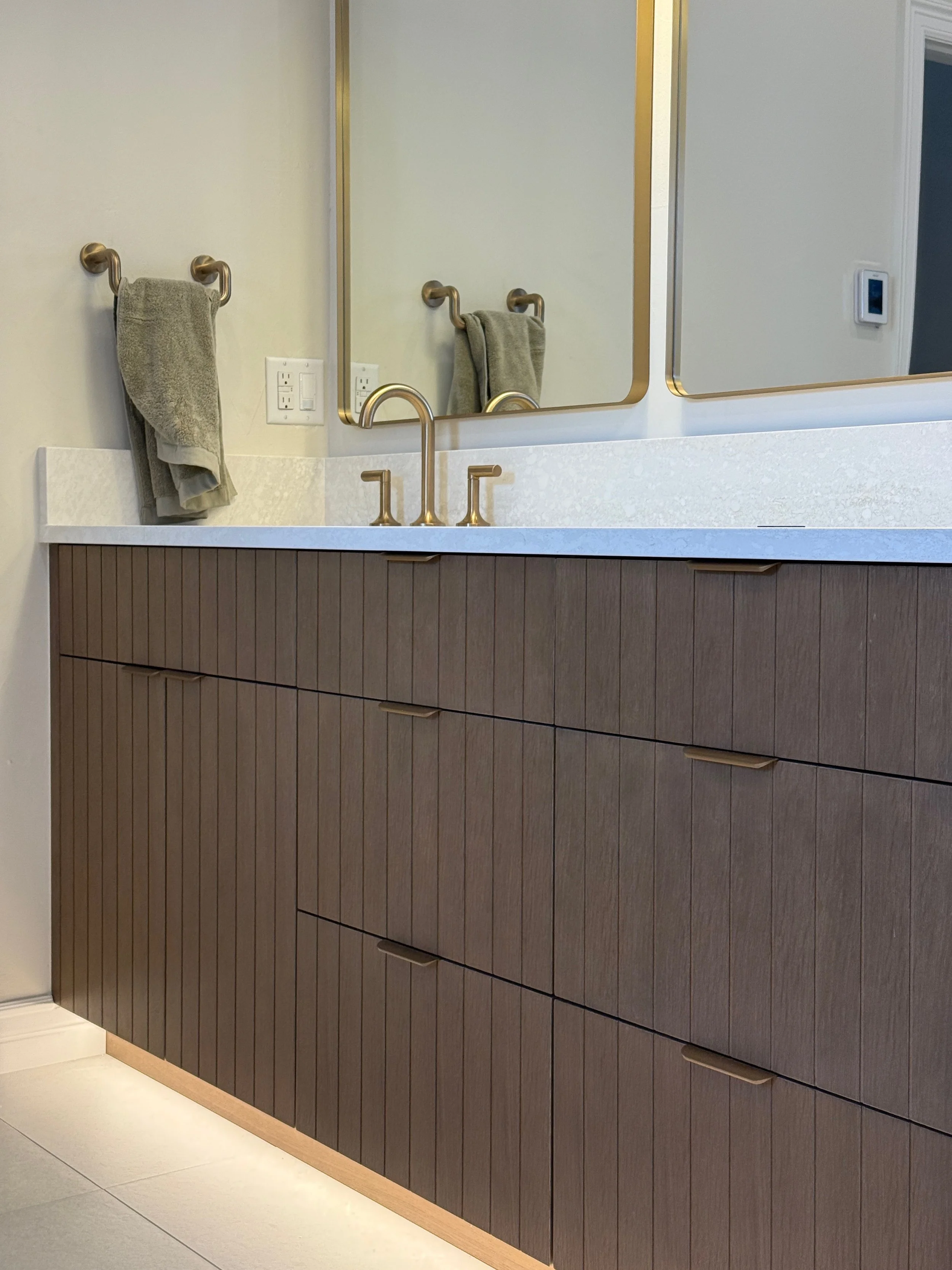 Modern bathroom vanity with wood cabinet drawers, a white countertop, a gold faucet, and a large mirror. Two beige towels hang on gold towel rings above the countertop.