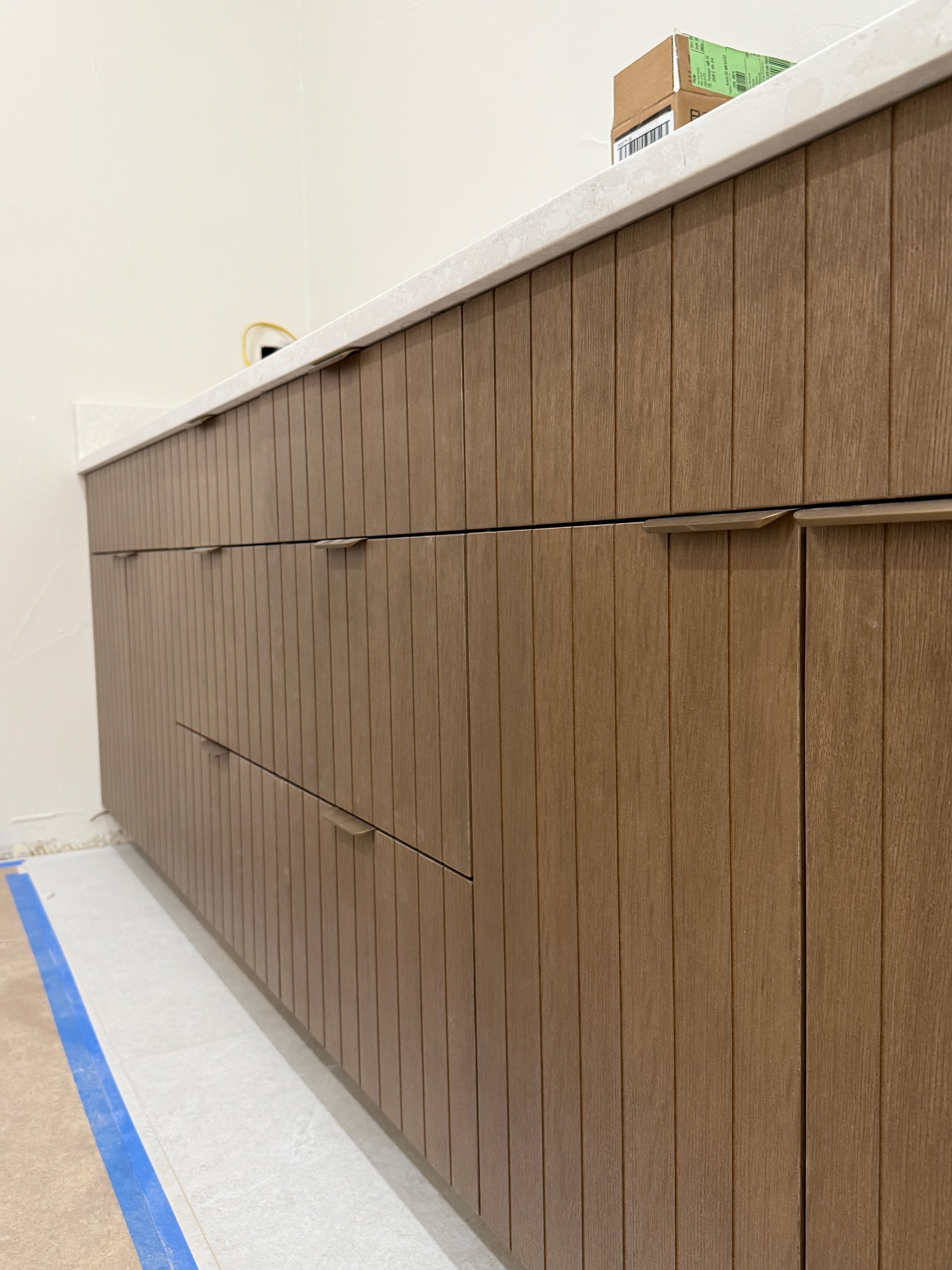 Brown wooden cabinet with vertical slats and a white countertop, with a cardboard box on top, located in an indoor setting.