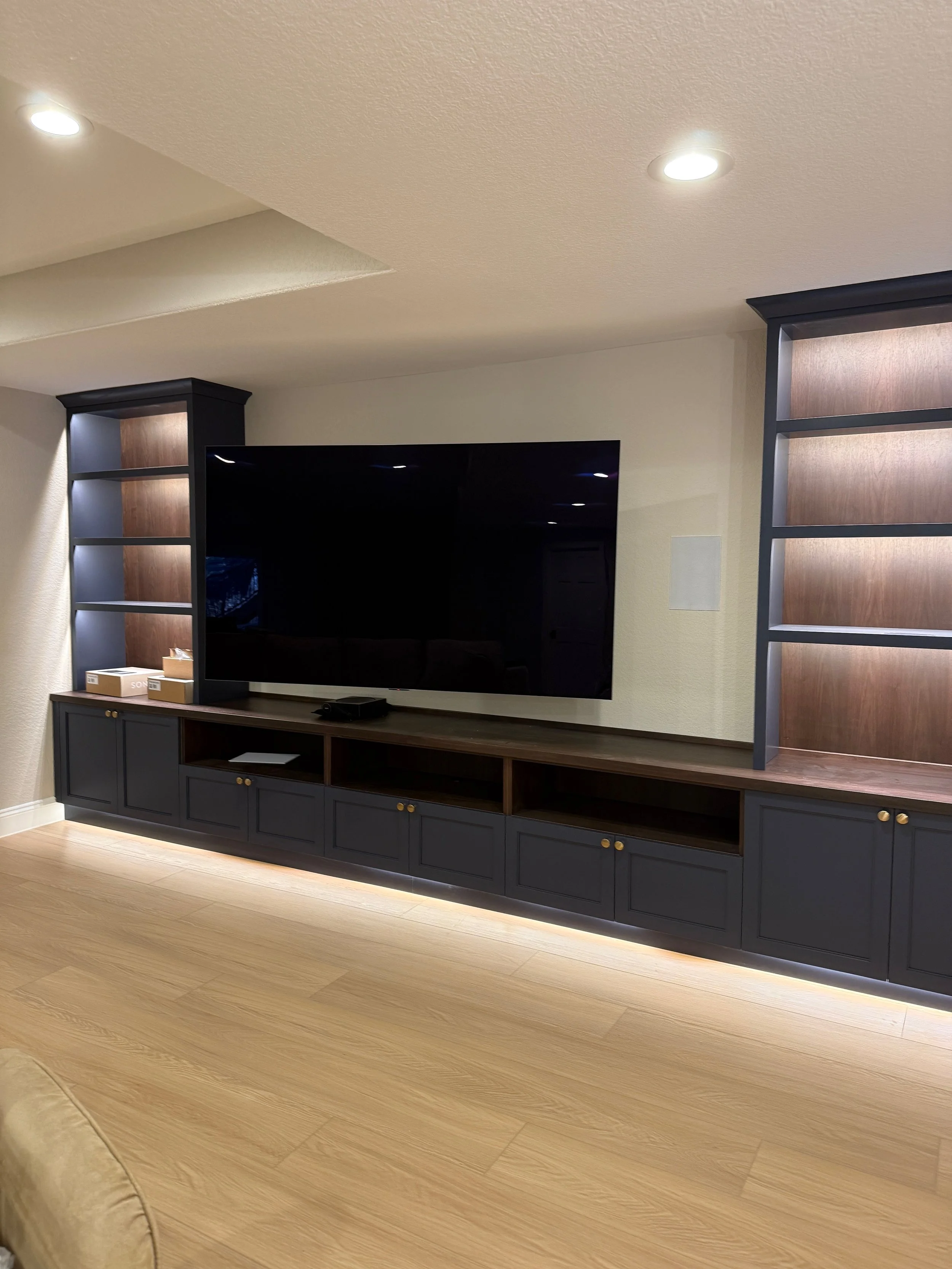 Modern living room entertainment center with large flat-screen TV, black and wood finished shelving and cabinetry, recessed lighting, and light wood flooring.